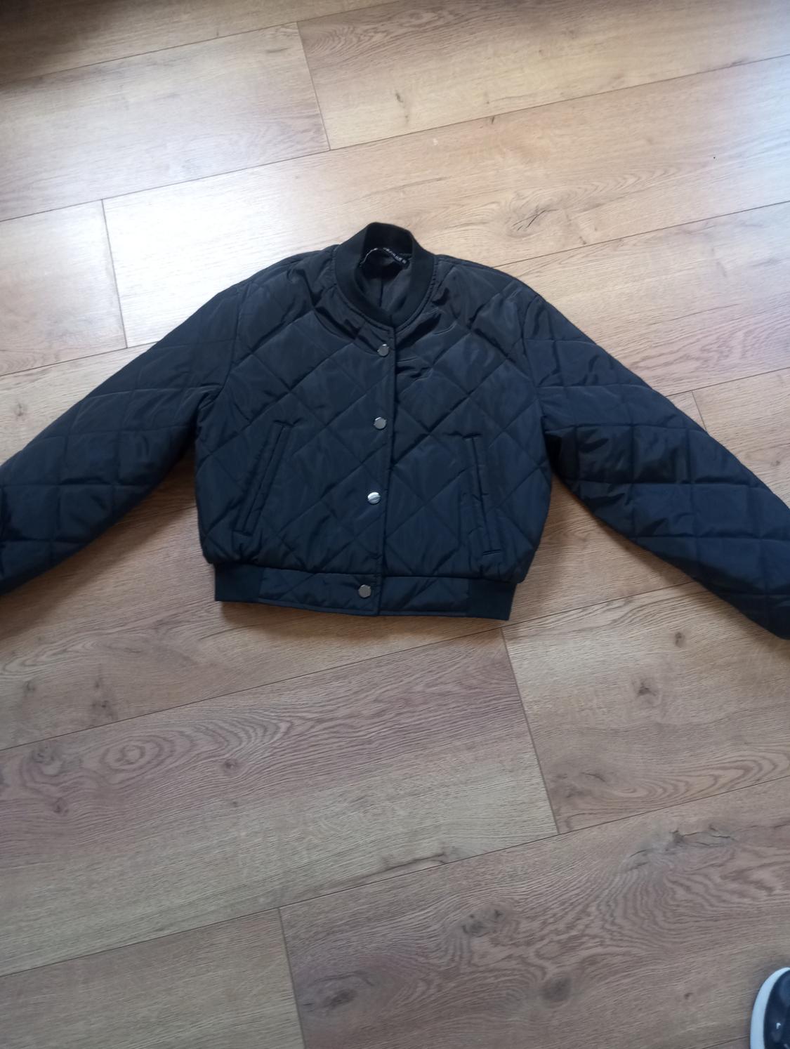 Black quilted jacket with snap fasteners, on a light wooden floor. Extended sleeves, casual style.