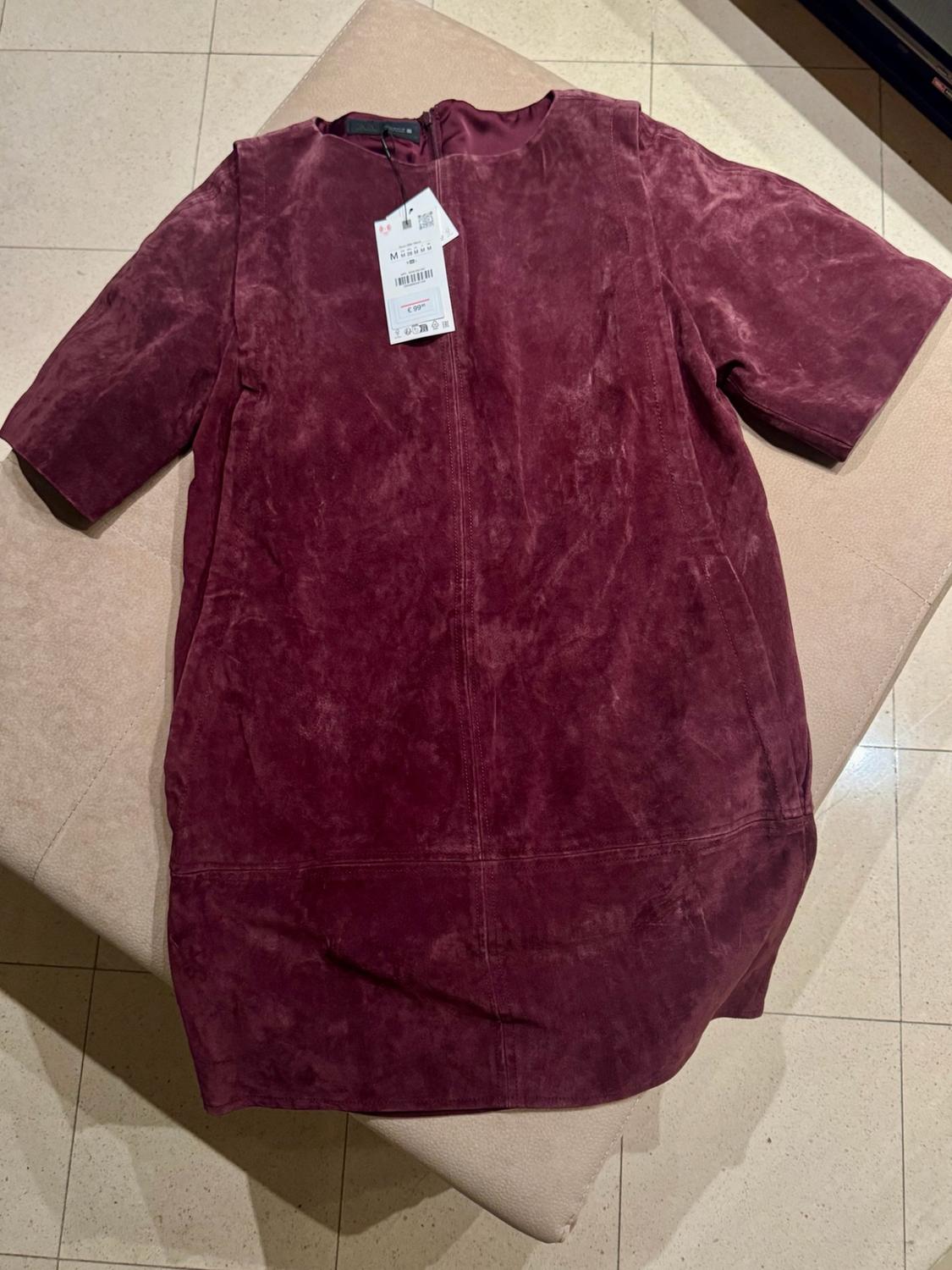 Short-sleeved burgundy dress with a suede texture and visible stitching, with an M label and a price of €99, on a beige seat.