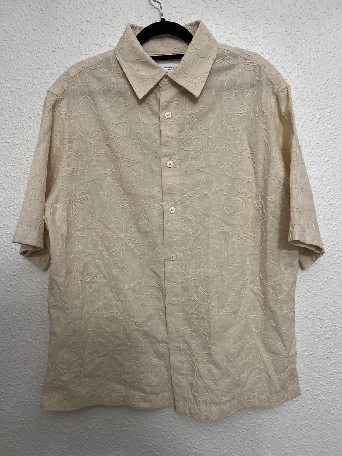 Beige short-sleeved shirt with floral embroidery hanging on a black hanger against a textured white wall.