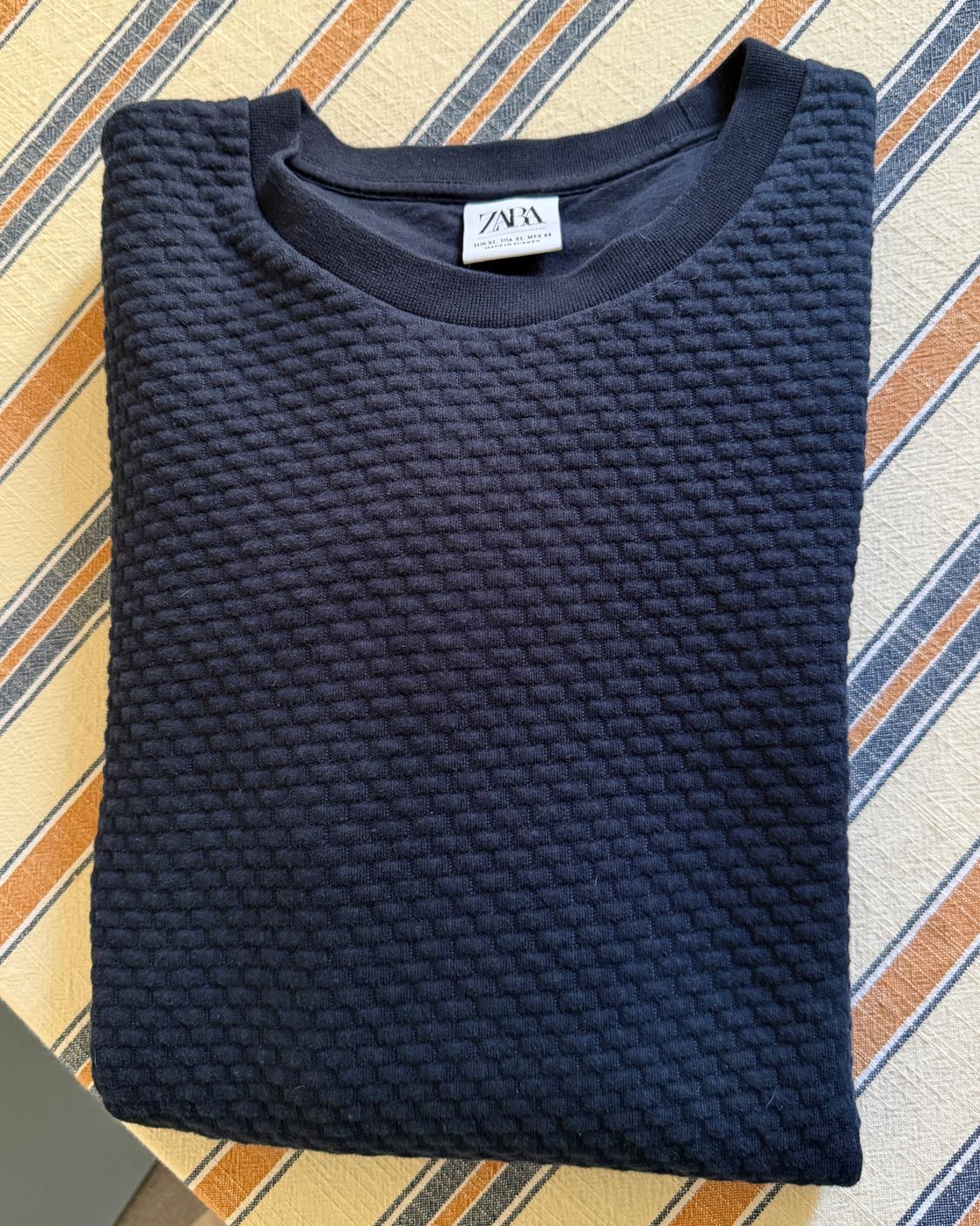 Navy blue sweatshirt with embossed texture, folded over a striped fabric. Visible tag with the text ZARA EUR XL USA XL MEX 44.