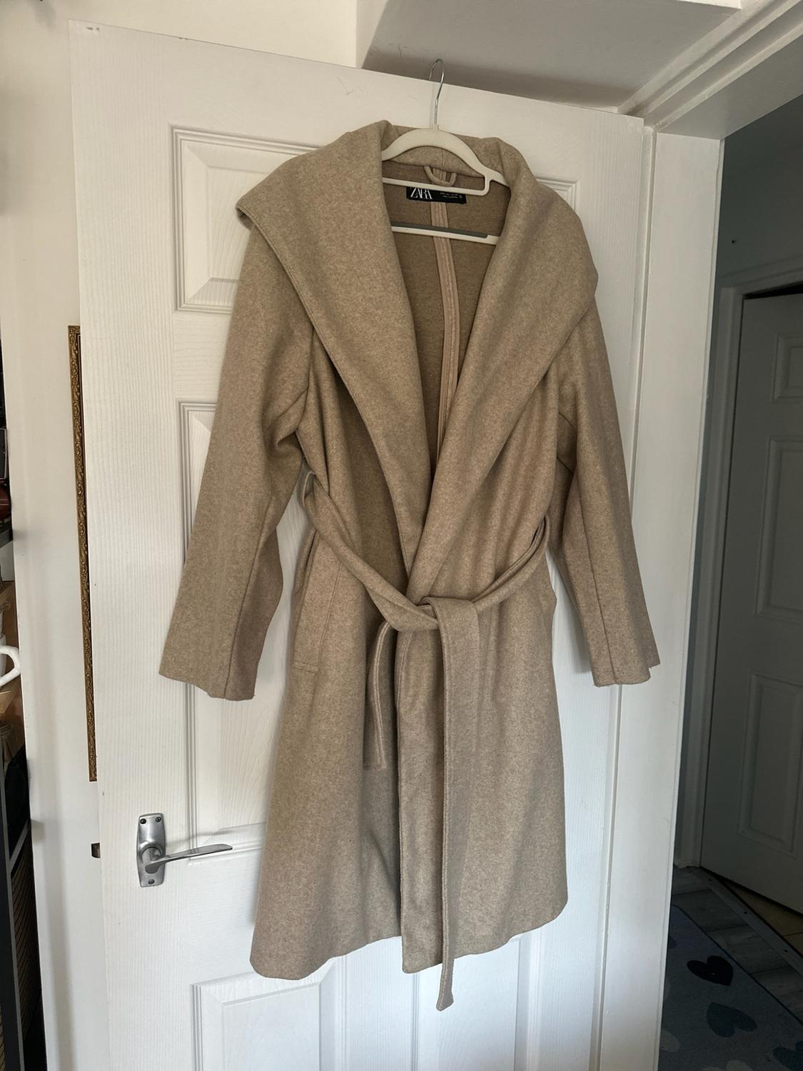 Beige coat with a hood and tied belt, hanging on a white door in a homely setting. The label reads ZARA.