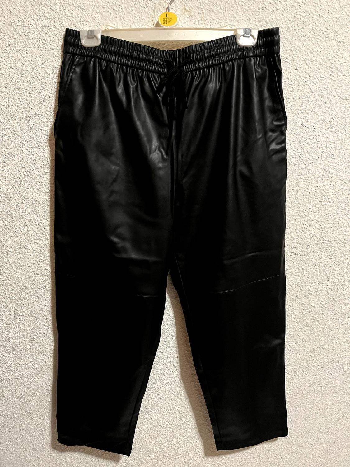Black trousers hanging on a white hanger with a yellow size L tag, against a white textured wall.