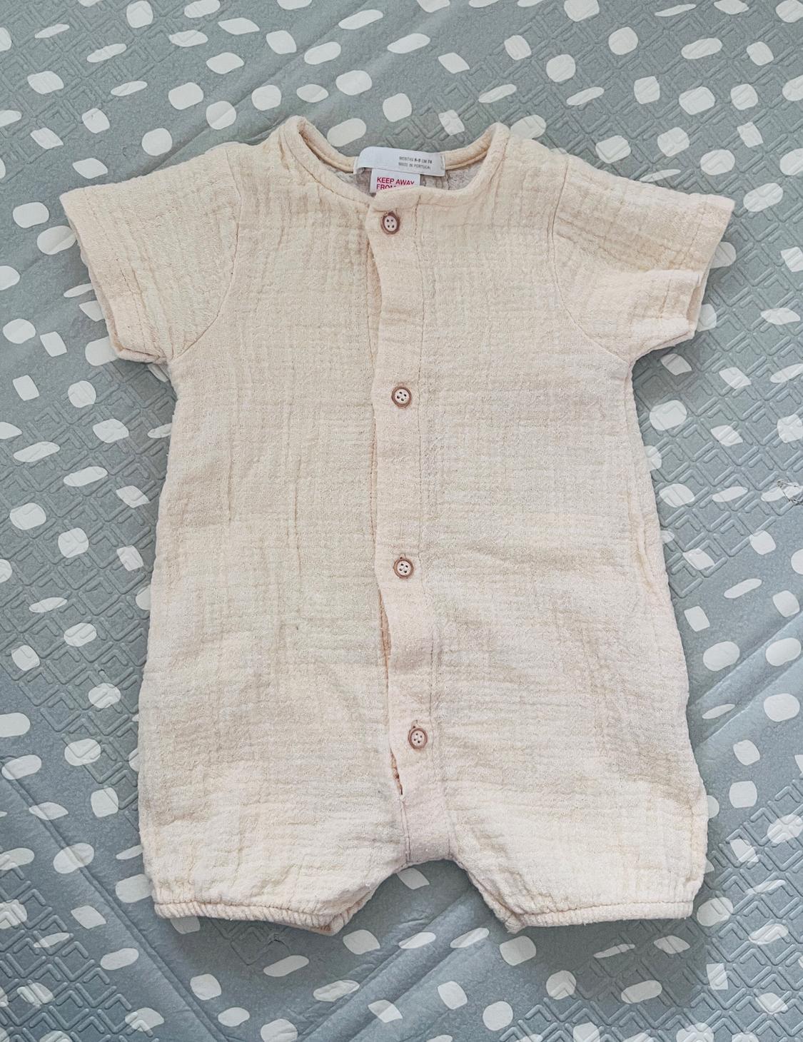Cream babygrow with front buttons, on a grey blanket with white patterns. Visible label: "KEEP AWAY FROM FIRE".