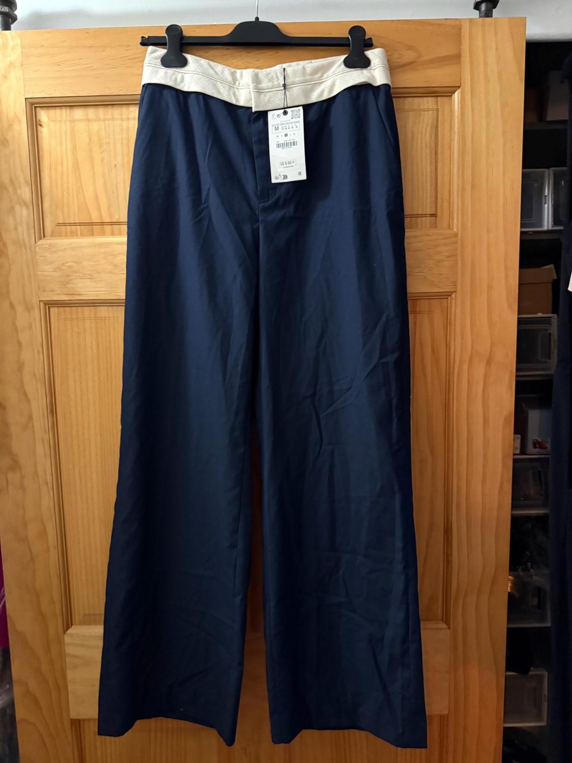 Wide navy blue trousers with a white waistband, hanging on a black hanger over a wooden door.
