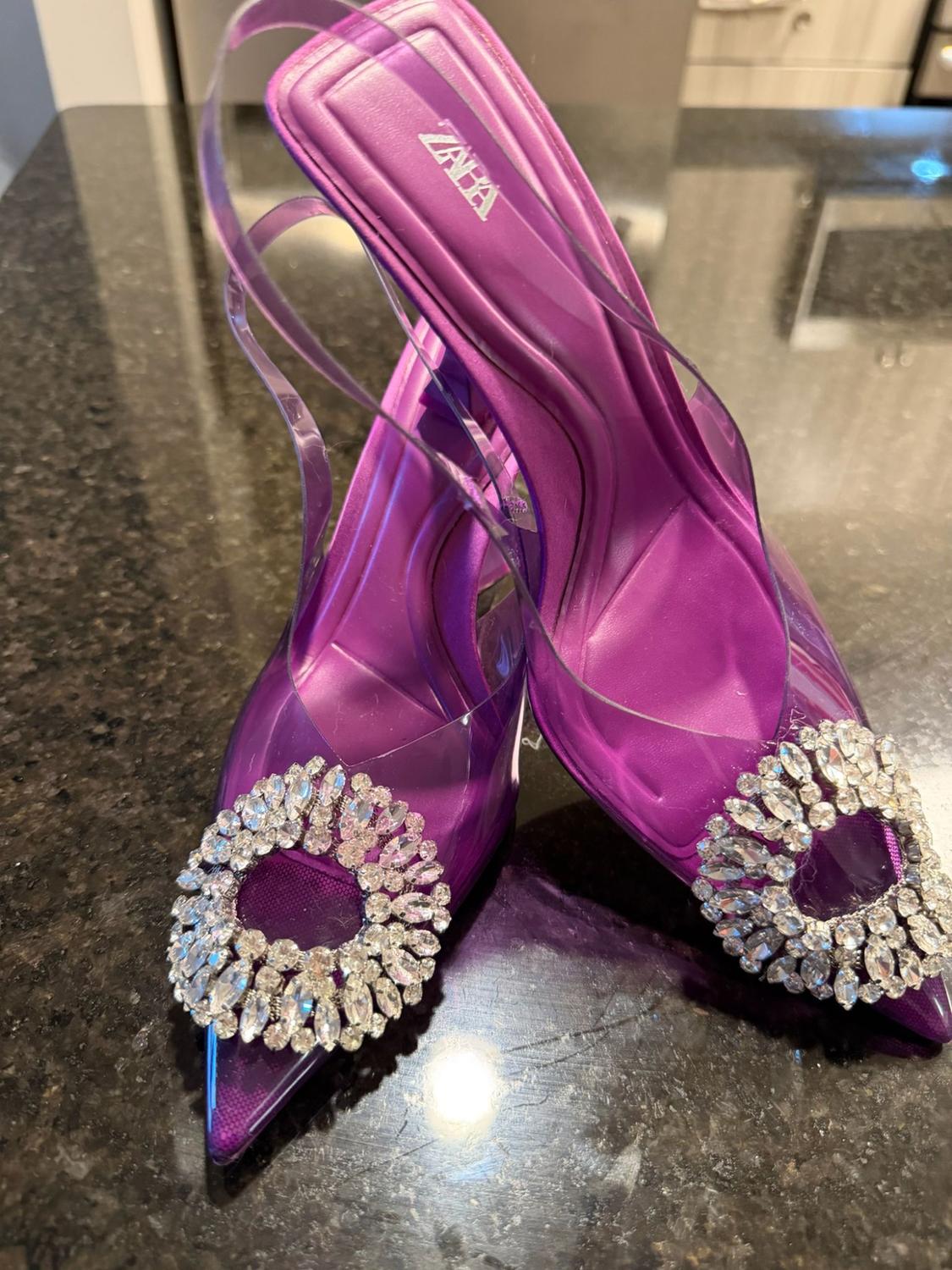 Purple high heels with a transparent toe and a circular rhinestone embellishment on a dark surface. Text: ZARA.
