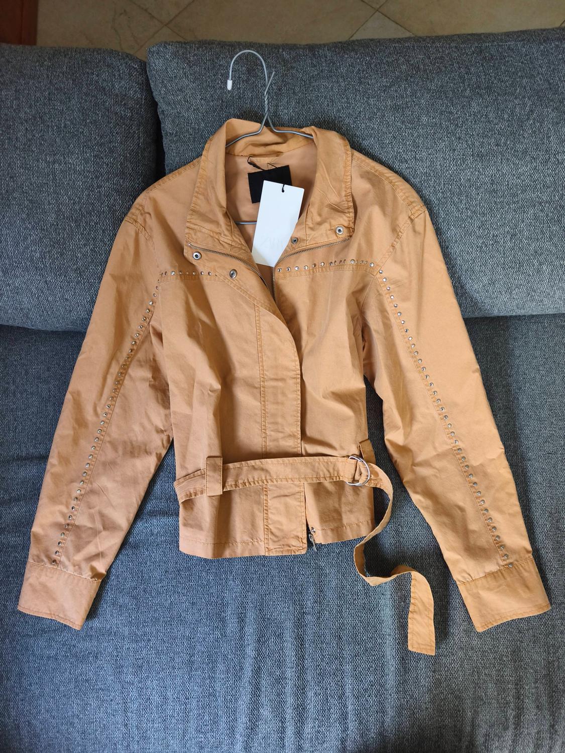 Mustard jacket with studs on the sleeves and a belt, hanging on a hanger over a grey sofa.