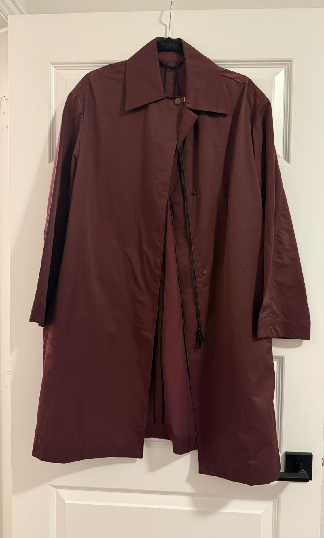 Long burgundy coat with a collar and zip, hanging on a hanger with the text 'kensie' above a white panelled door. Casual style.