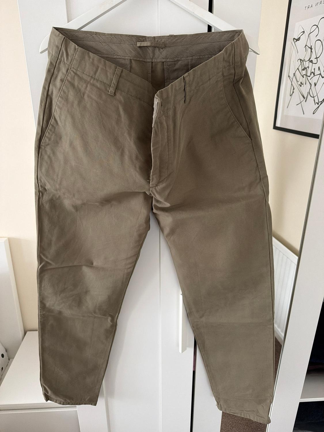 Khaki trousers hanging on a white hanger in front of a white wardrobe, with an abstract painting on the wall in the background.
