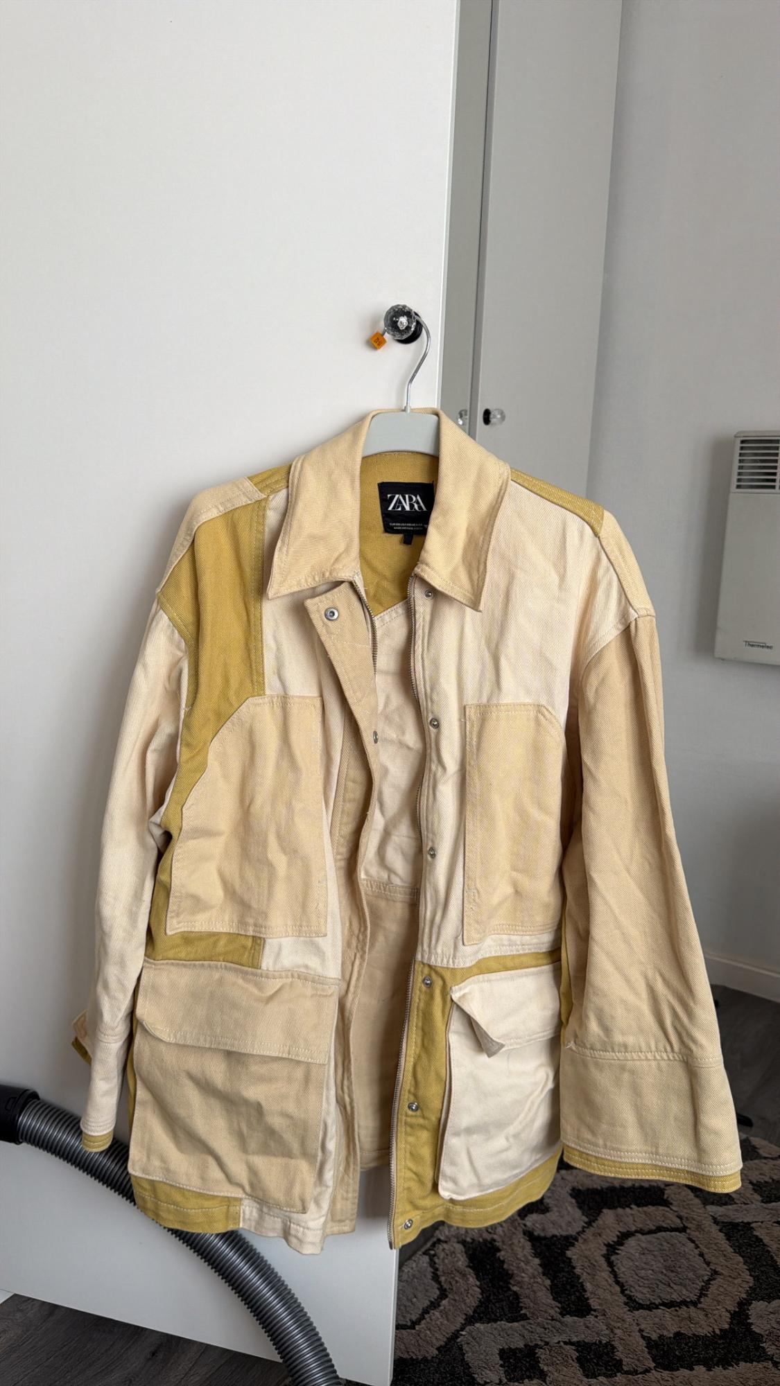 Beige and mustard panelled jacket with pockets hanging on a white door. Relaxed atmosphere. Label with the text ZARA.