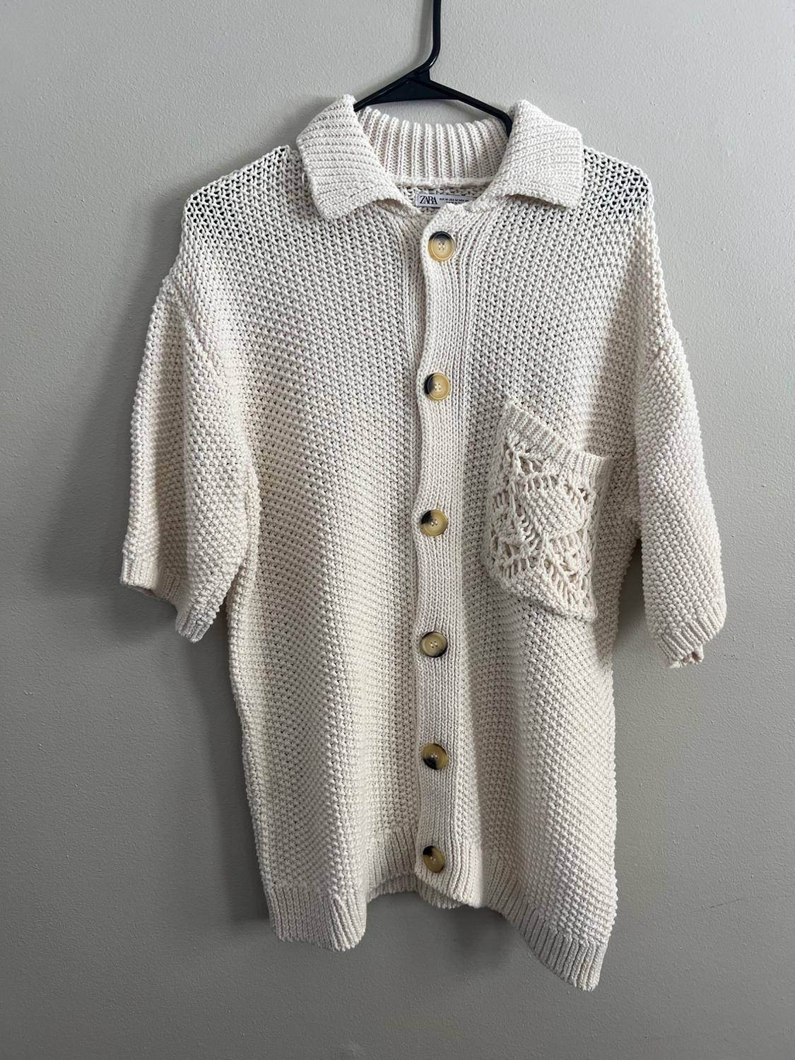 White knit cardigan with large buttons and a woven front pocket, hanging on a black hanger.