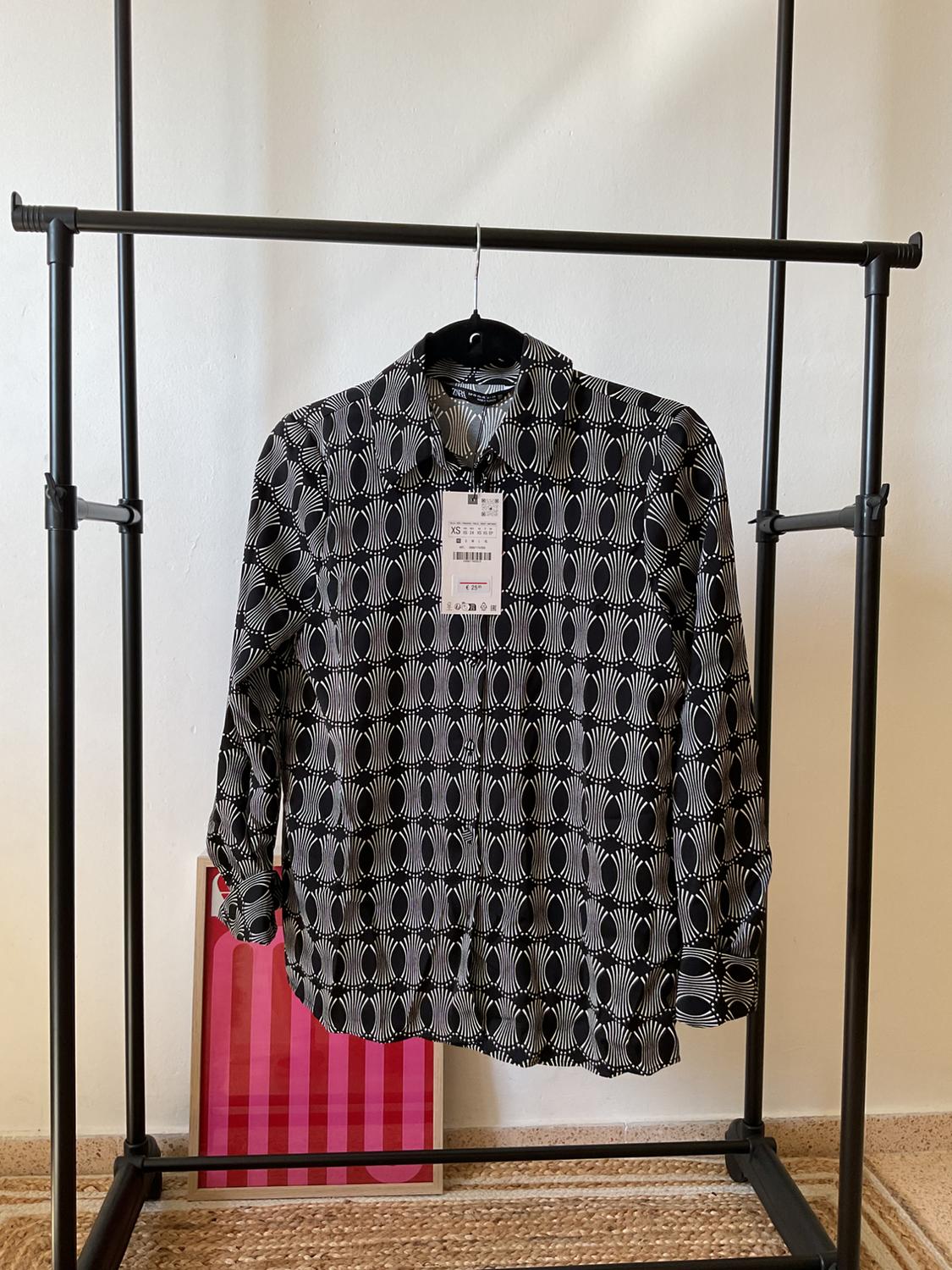 Shirt hanging on a black hanger, with a black and white geometric print. Visible label. Background with white wall and pink picture.