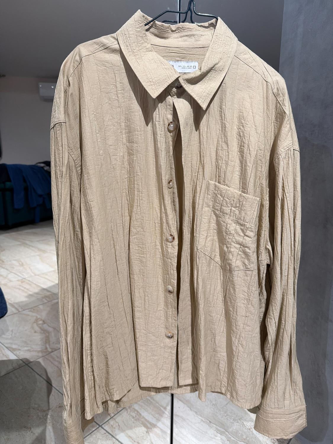 Beige crinkle-fabric shirt hanging on a hanger, with front buttons and a chest pocket, in an indoor setting.