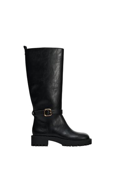 Black high boot with gold buckle and chunky sole. White background.