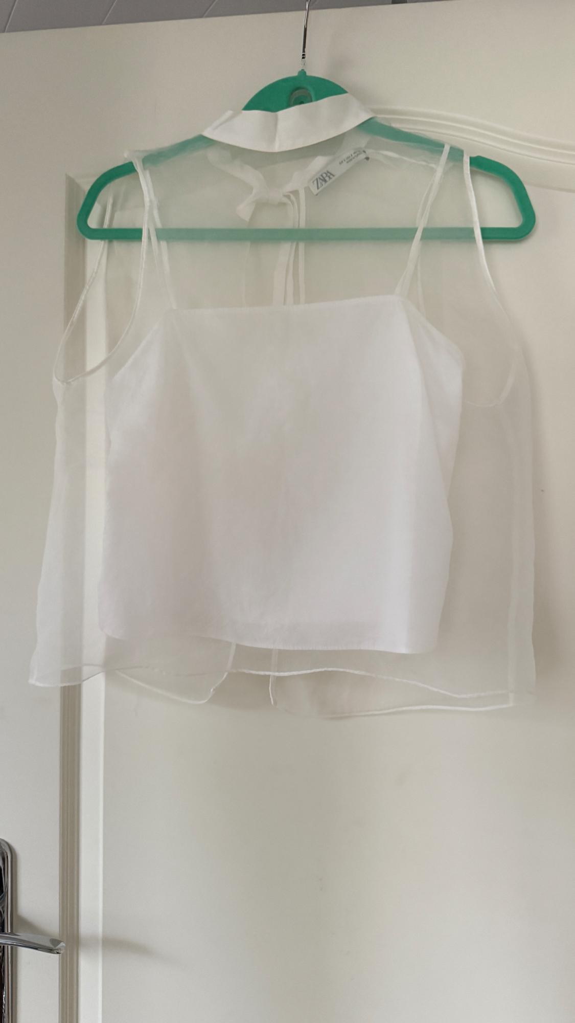 Sleeveless white blouse made of sheer fabric hanging on a green hanger. Visible tag with "ZARA" text. White door background.