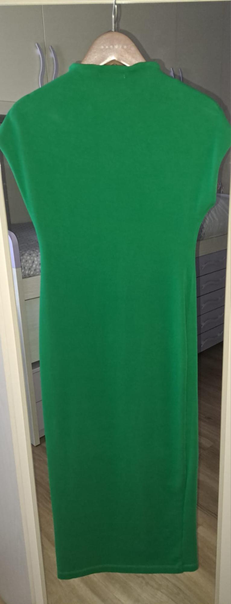 Long green sleeveless high-neck dress hanging on a wooden hanger. In the background, a bedroom with a bed is visible.