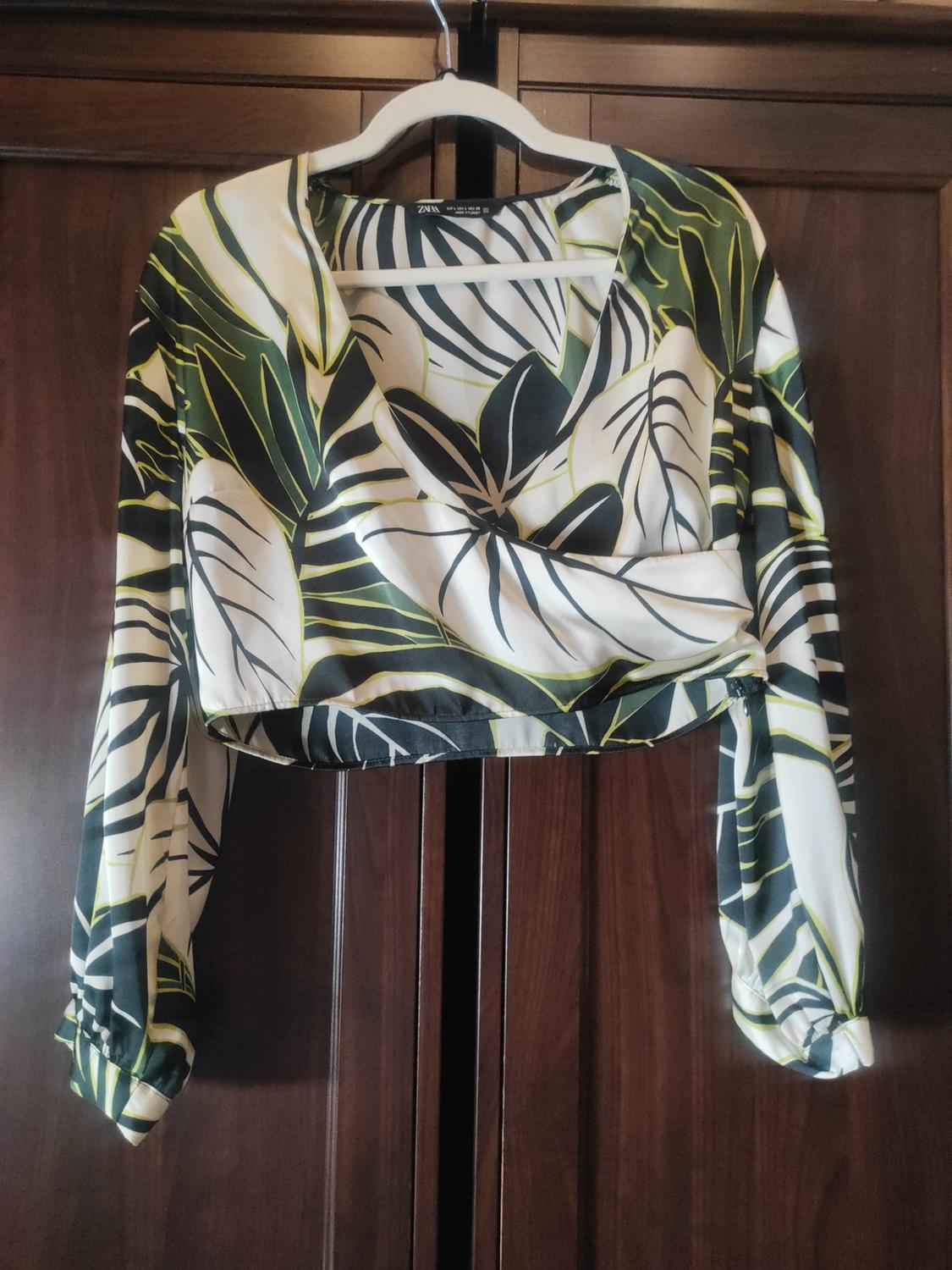 Cropped long-sleeved wrap blouse with a tropical leaf print, hanging in a dark wooden wardrobe. ZARA text.