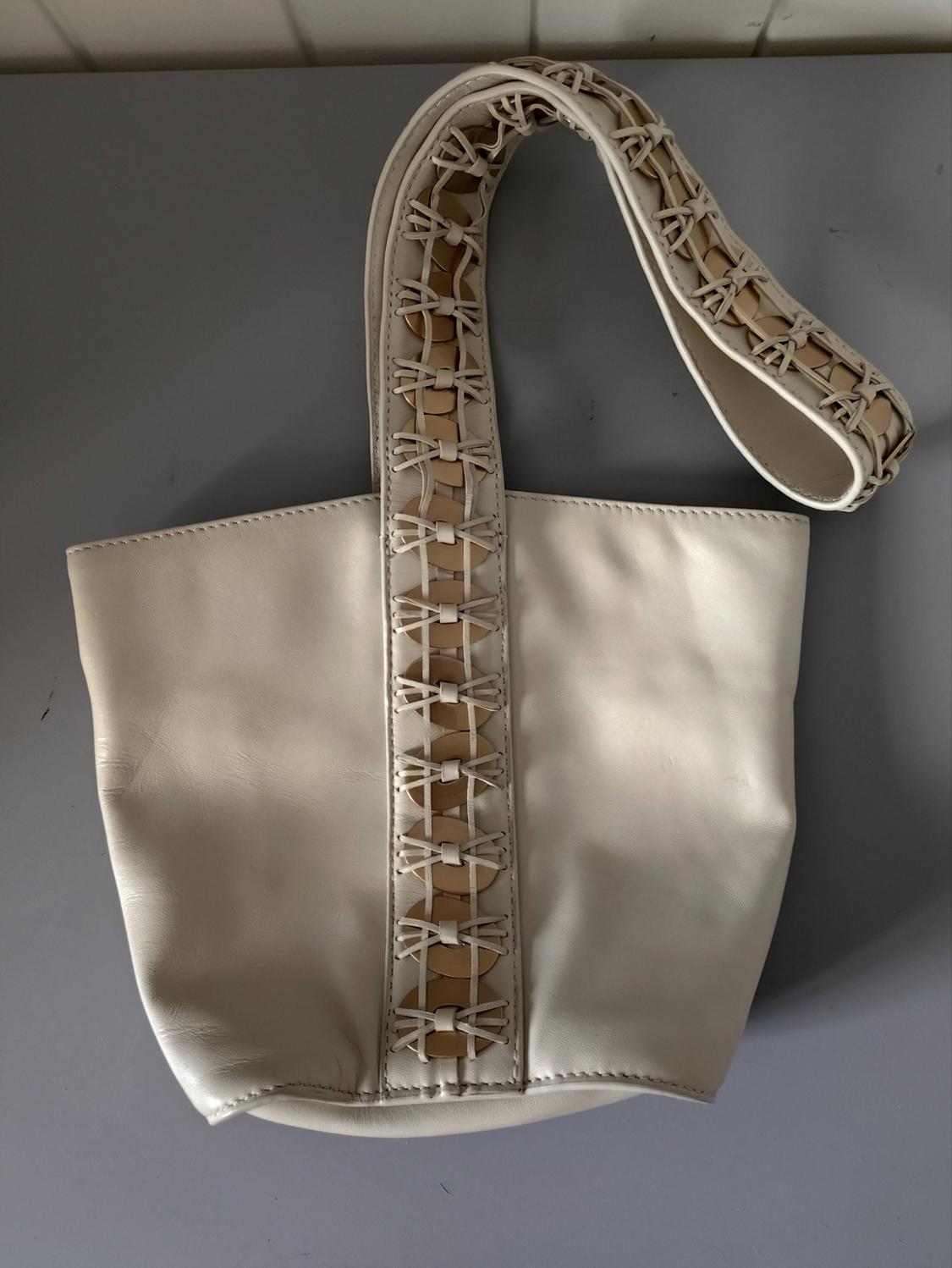 Cream leather bag on a grey surface. Its central handle with a braided design and circular details in a beige tone stands out.