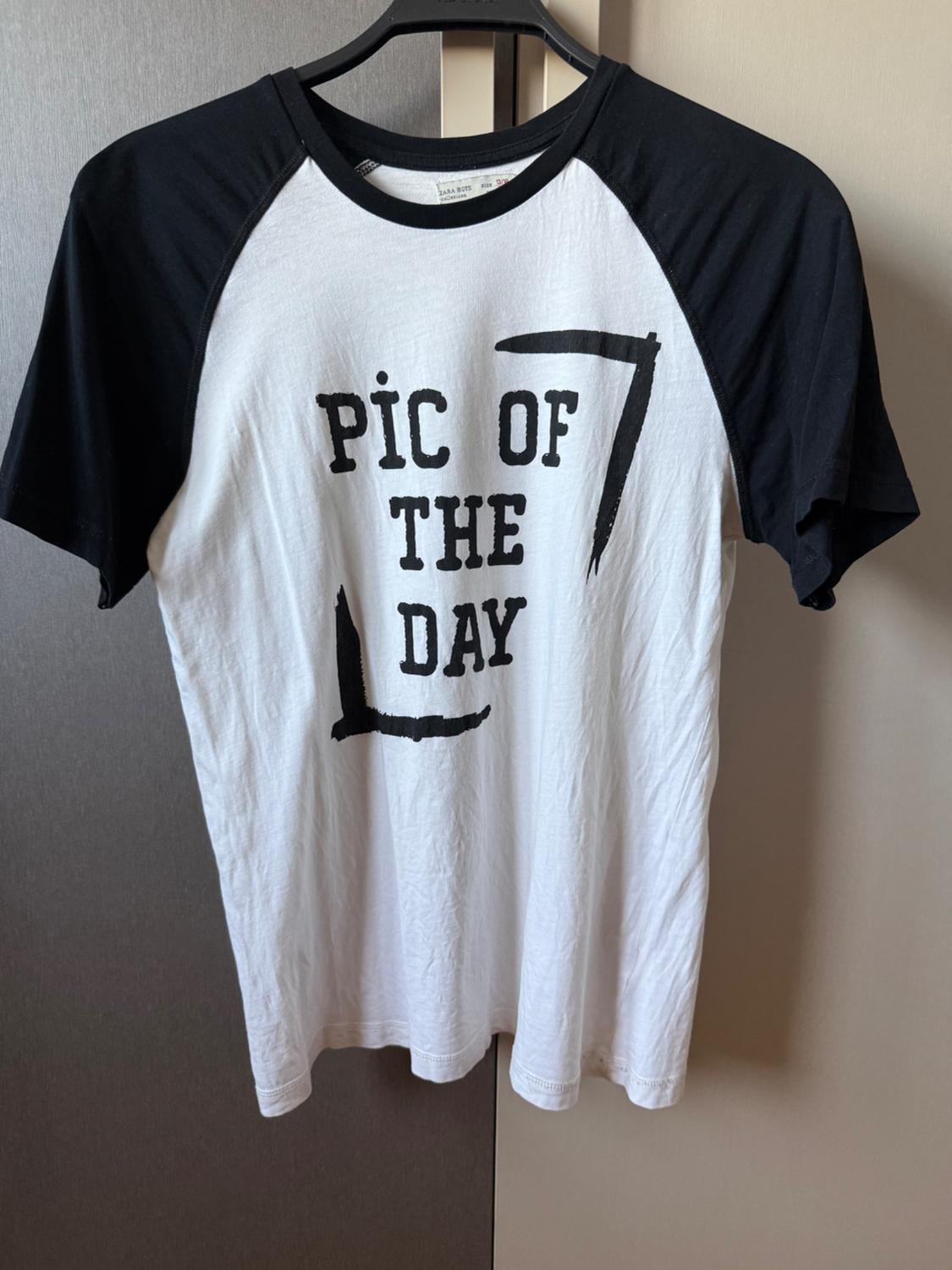 White T-shirt with black sleeves hanging on a hanger. On the front, it has the text "PIC OF THE DAY" printed in black.