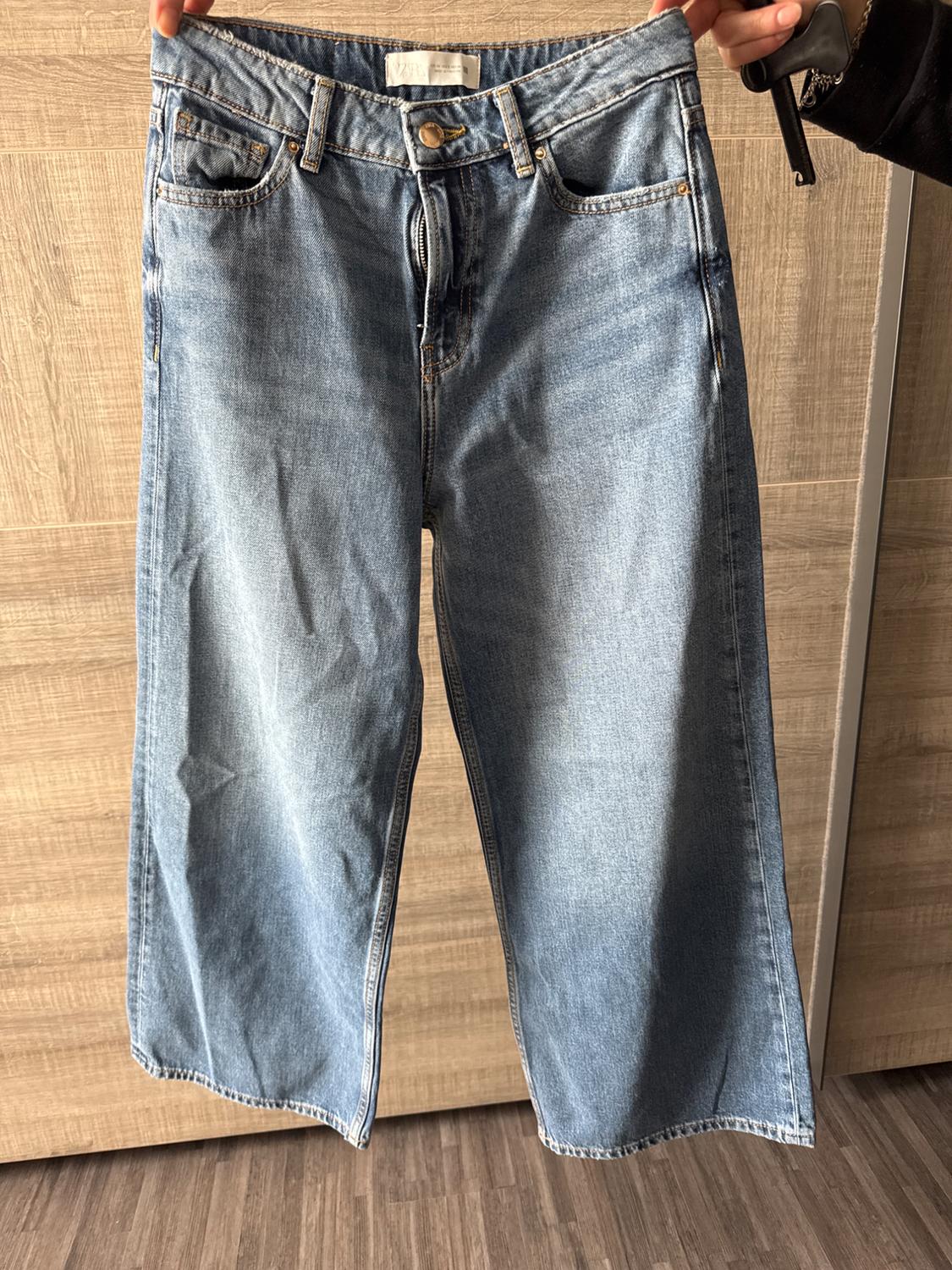 A pair of light blue denim jeans with a wide cut, held by a hand against a light wooden background.