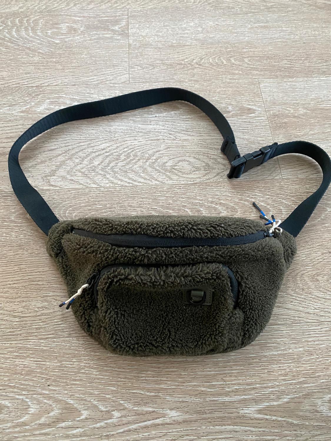 Brown plush fanny pack with black zipper closure and adjustable strap on a light wood floor.