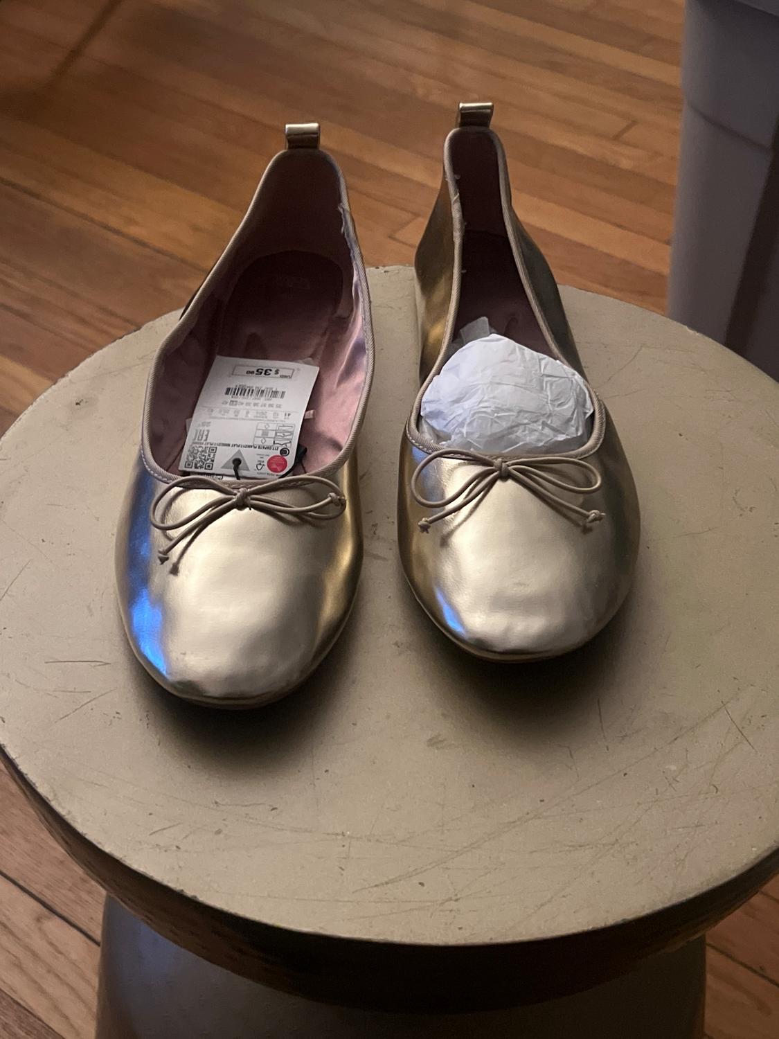 Gold shoes with a bow on a round wooden table. One has a tag and the other paper inside. Wooden floor background.
