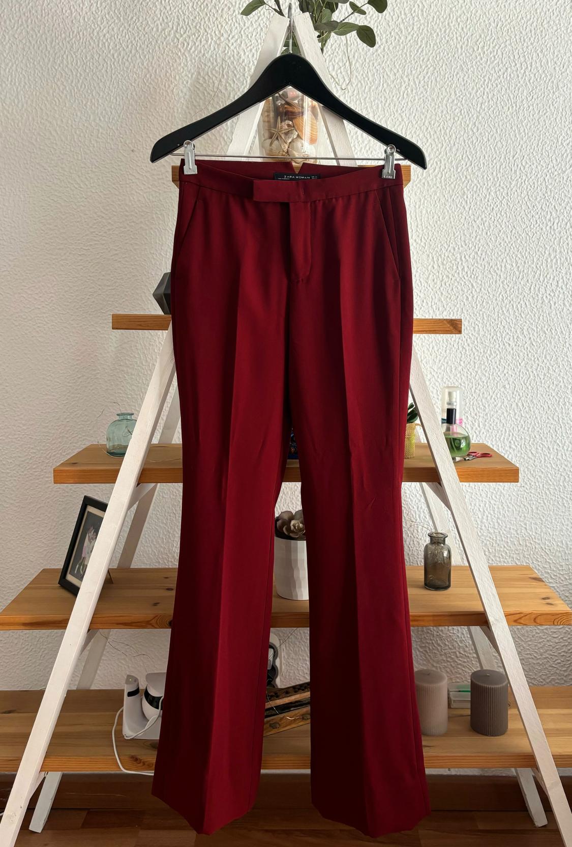 Red trousers hanging on a black hanger in front of a wooden bookshelf with decorative objects and plants.