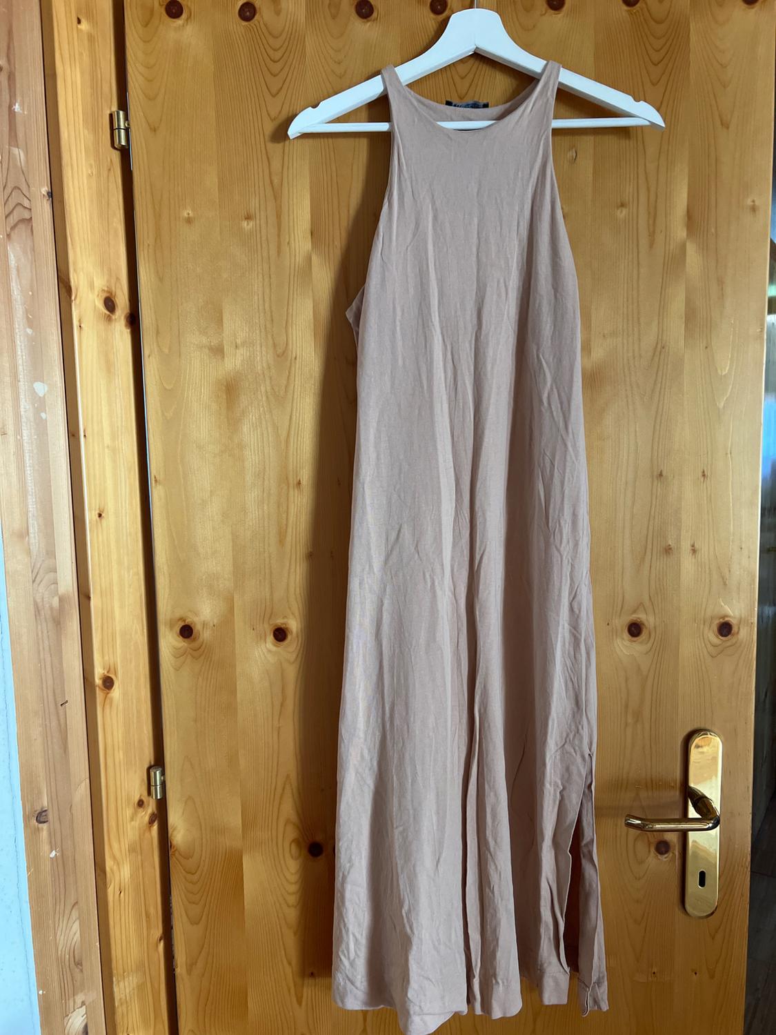Beige casual sleeveless maxi dress with a side slit, hanging on a white hanger against a light wood door.