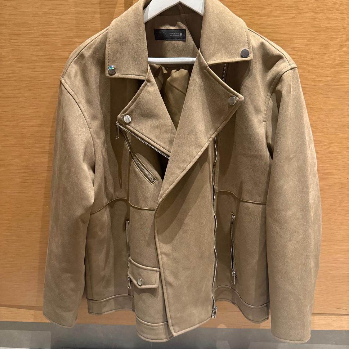 Beige biker-style jacket with zips and metal buttons, hanging on a hanger against a light wooden background.