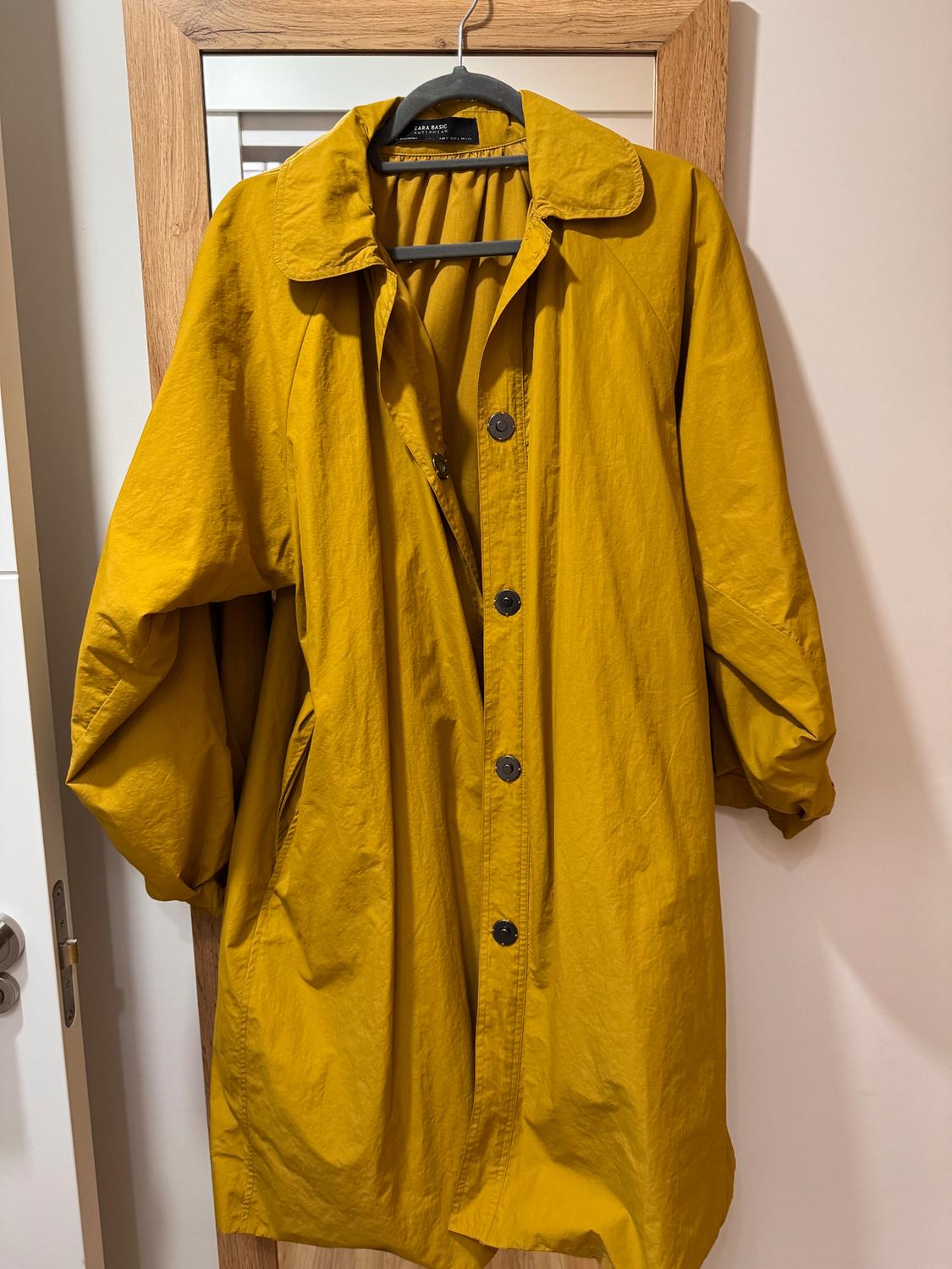 Mustard yellow coat hanging on a black hanger, with dark buttons and a light wood-framed mirror background.