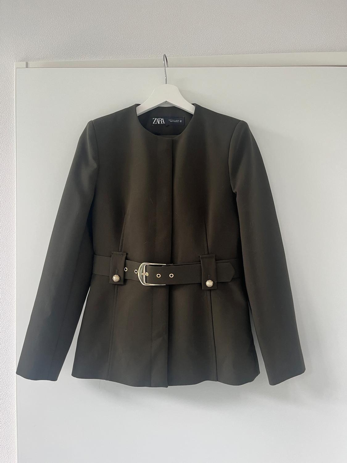 Olive green jacket with a belt and silver buckle, hanging on a white hanger. Visible label: Zara. White background.
