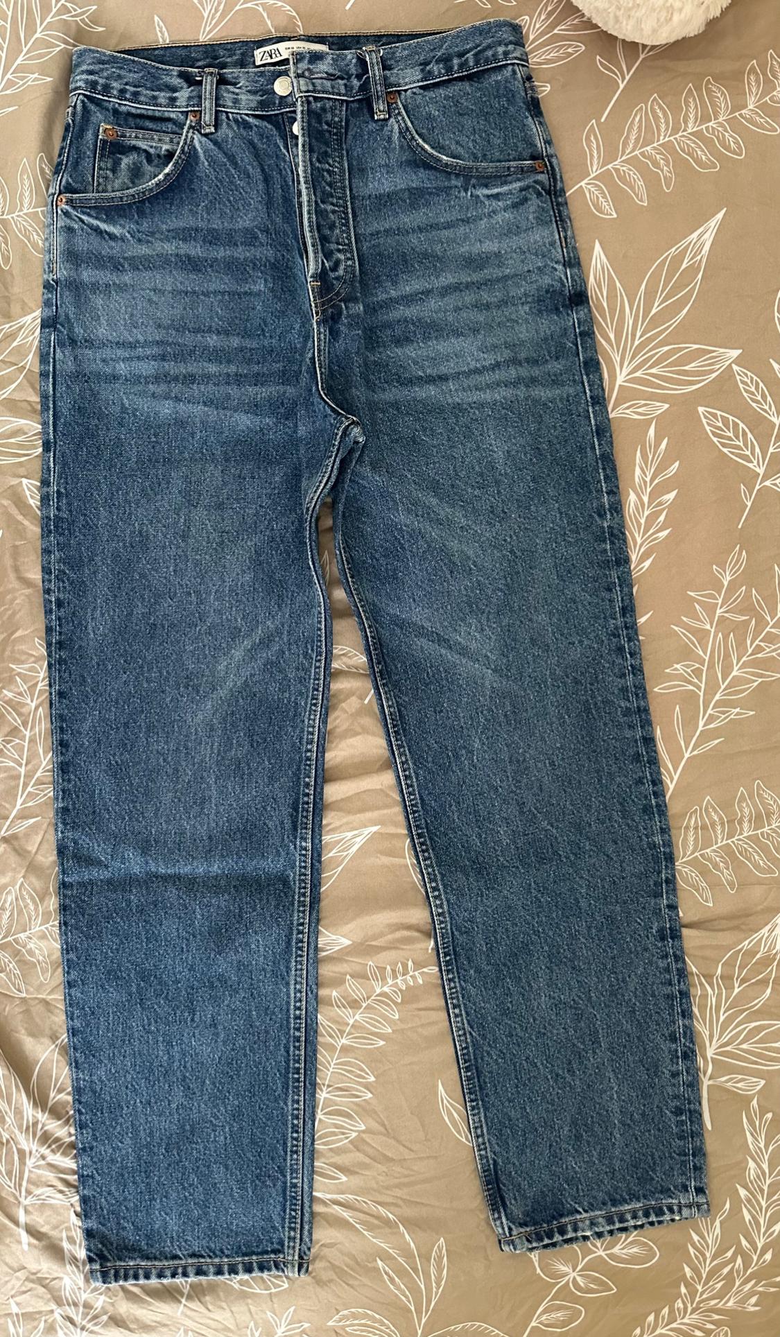 Blue denim jeans with a ZARA label, on beige fabric featuring a white leaf print. Casual style.