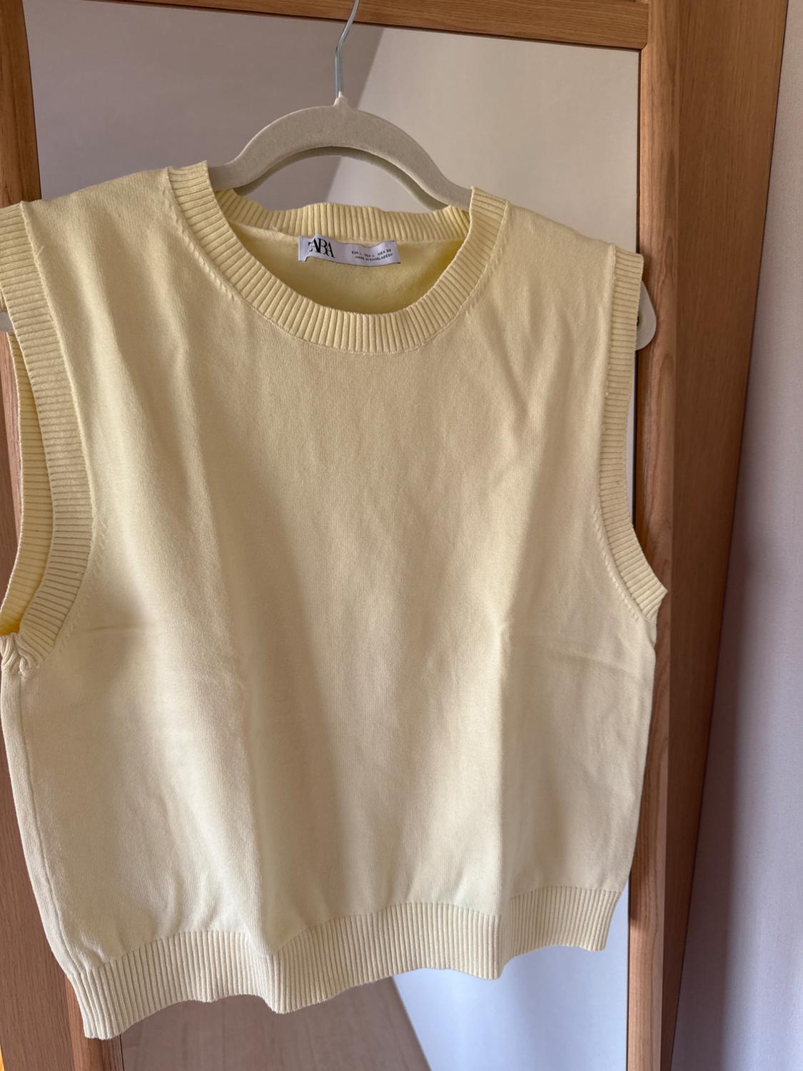 Cream sleeveless waistcoat hanging on a wooden hanger in front of a mirror. Label visible on the collar.