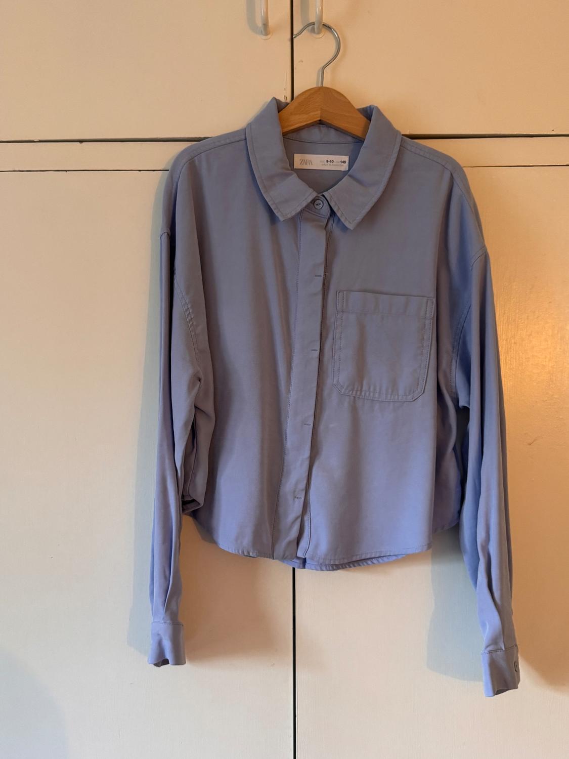 Light blue, long-sleeved shirt hanging on a wooden hanger against a white wardrobe background. Label visible on the collar.