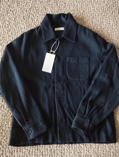 Black button-up jacket with a chest pocket and white label, on a beige rug.