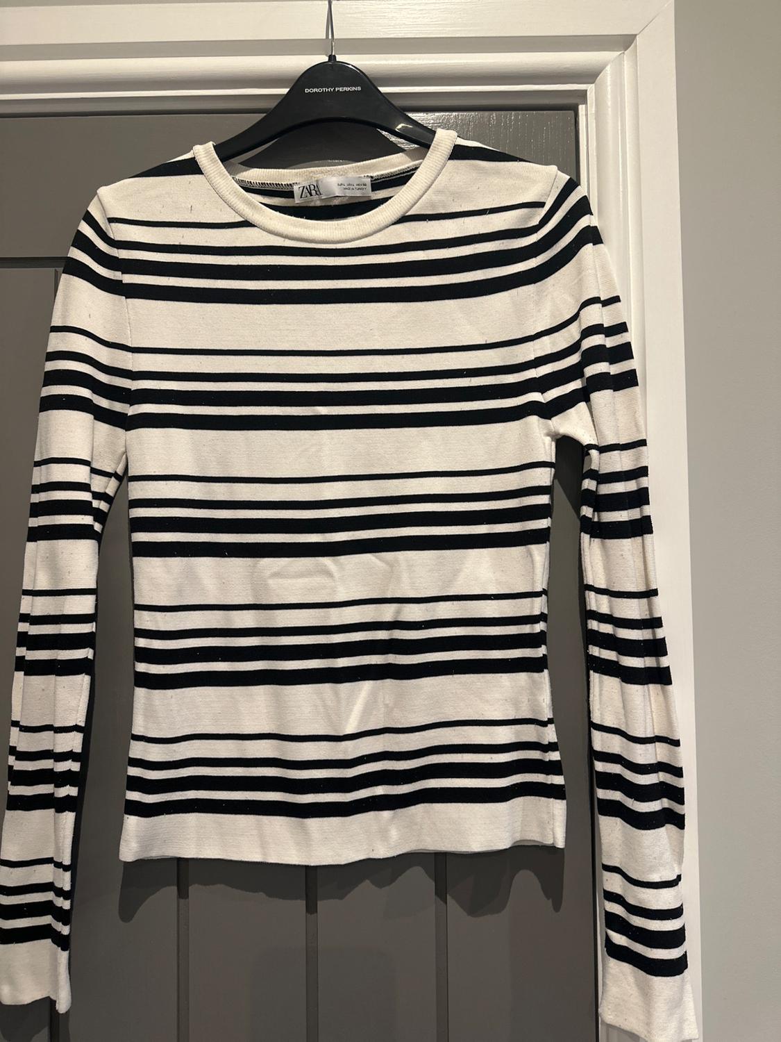 Black and white striped jumper hanging on a black coat hanger with the label visible. Grey door background and white frame.