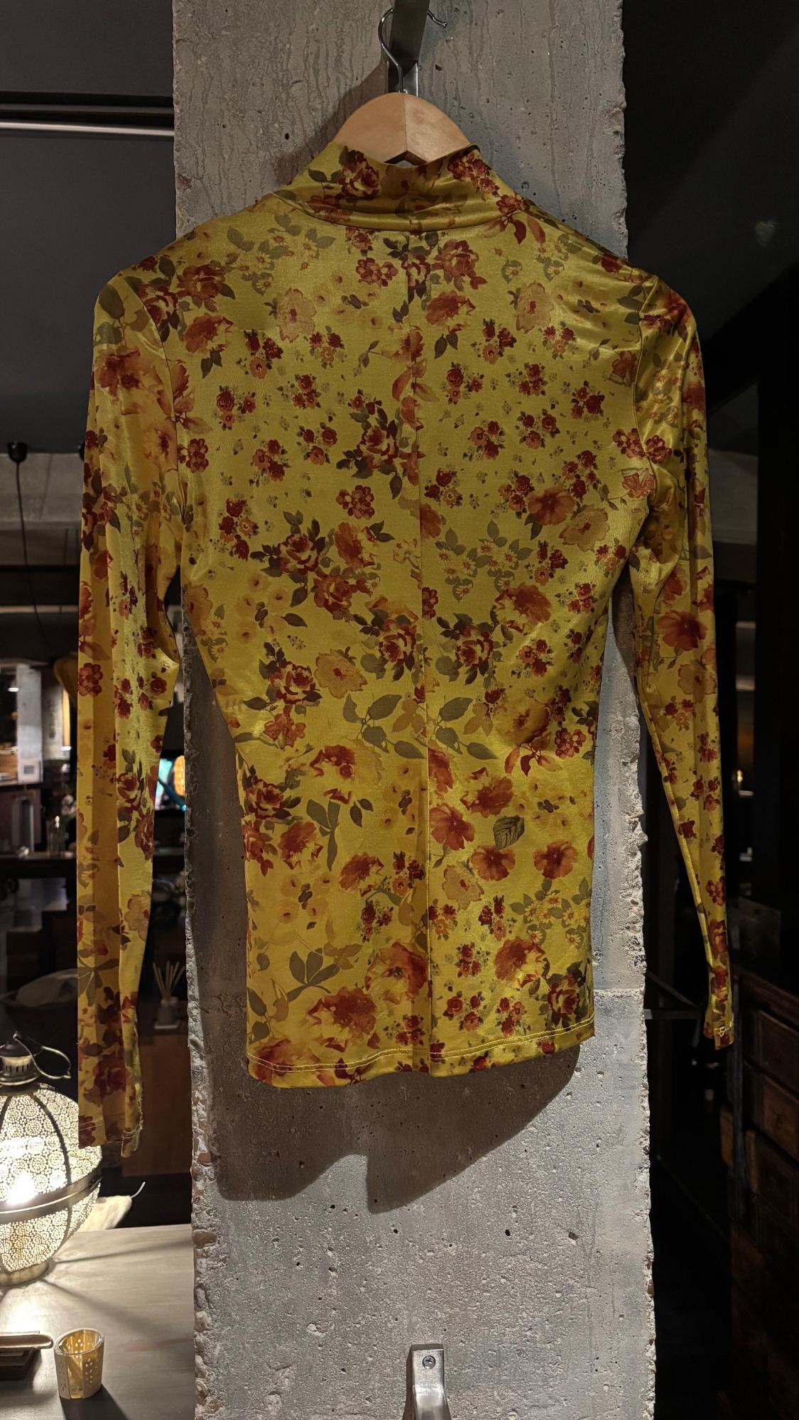 Yellow long-sleeved, high-neck floral print top, hanging on a wooden hanger against a concrete pillar indoors.