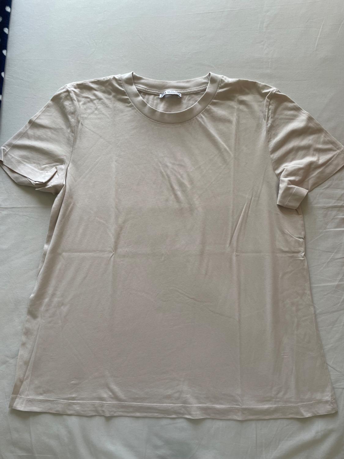 Beige short-sleeved, round-neck T-shirt on a light surface, with visible neck label.