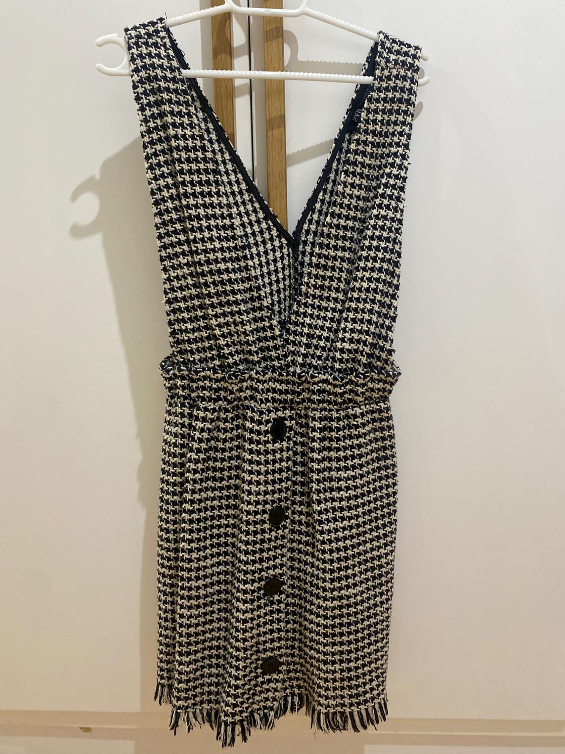 Sleeveless black and white checked dress, with front buttons and fringing on the hem, hanging on a white hanger.