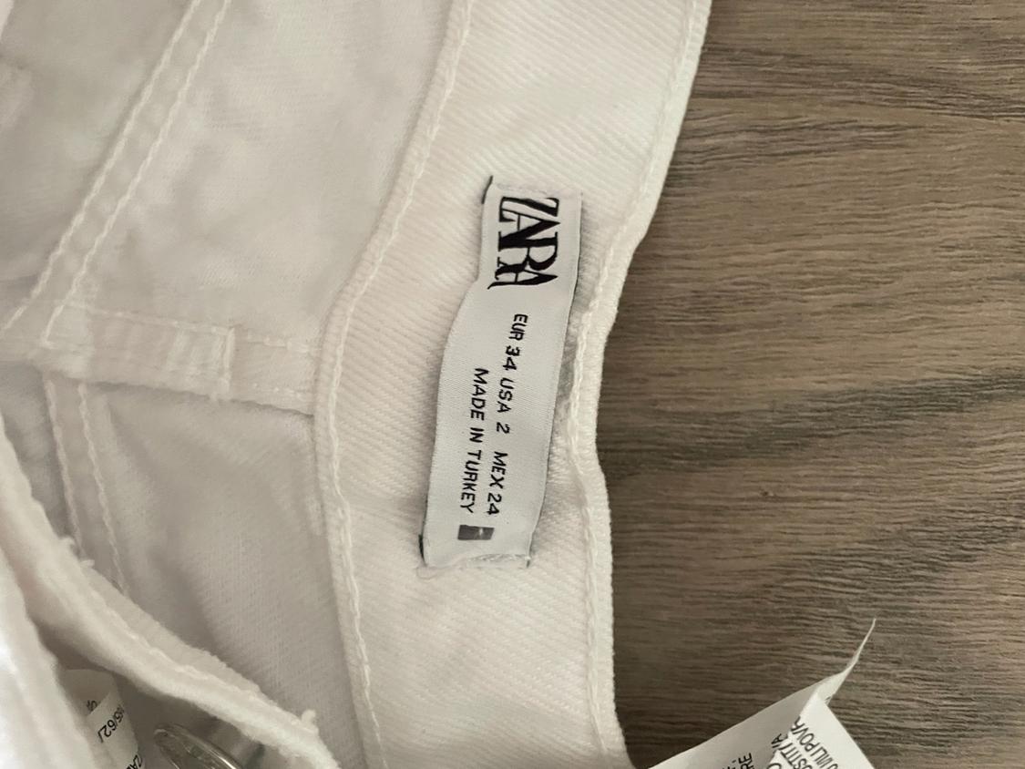 Detail of a white garment on wood with a tag showing the text "ZARA EUR 34 USA 2 MEX 24 MADE IN TURKEY".