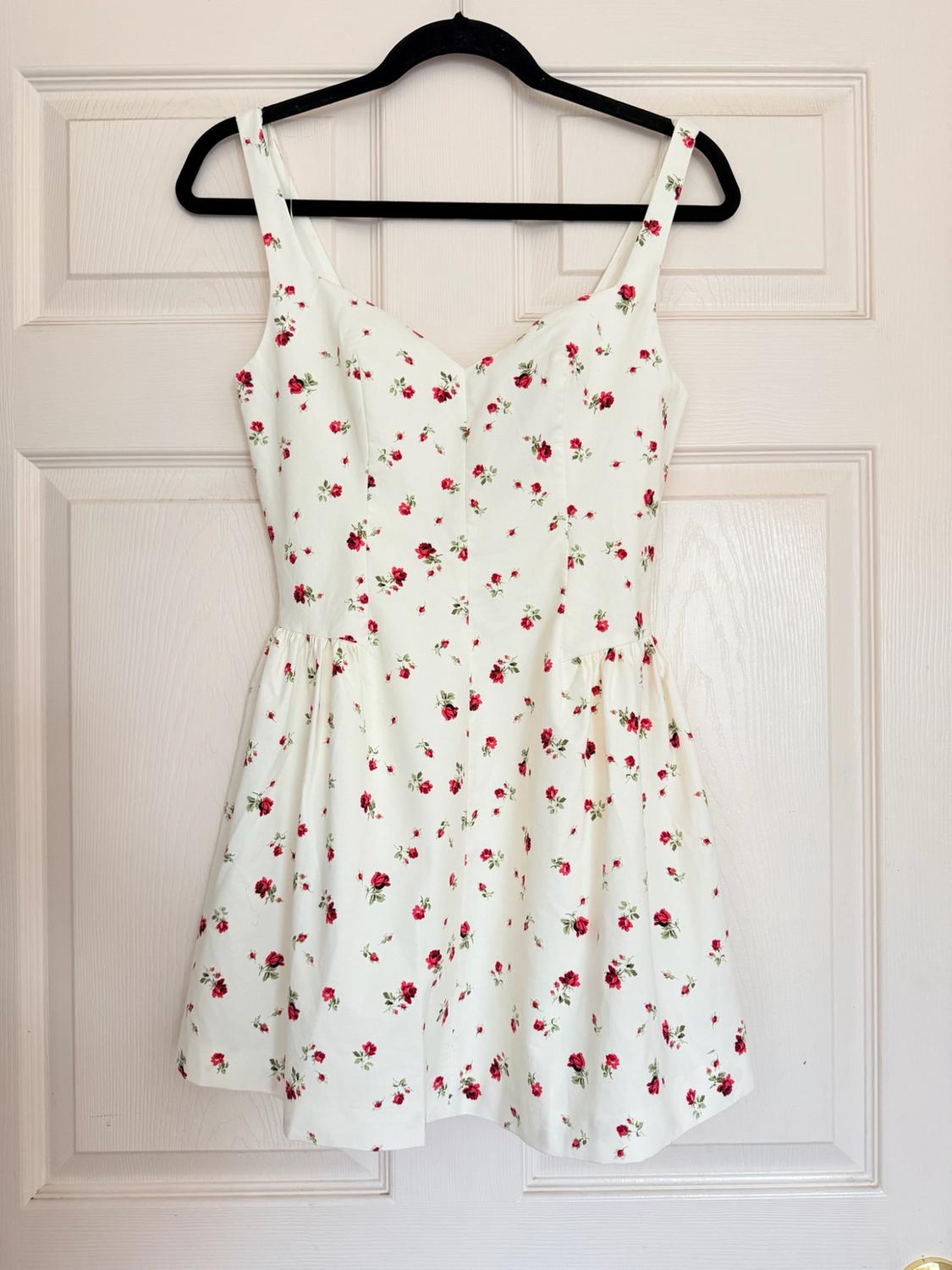 Short white dress with a pattern of small red flowers, hanging on a black hanger on a white door.