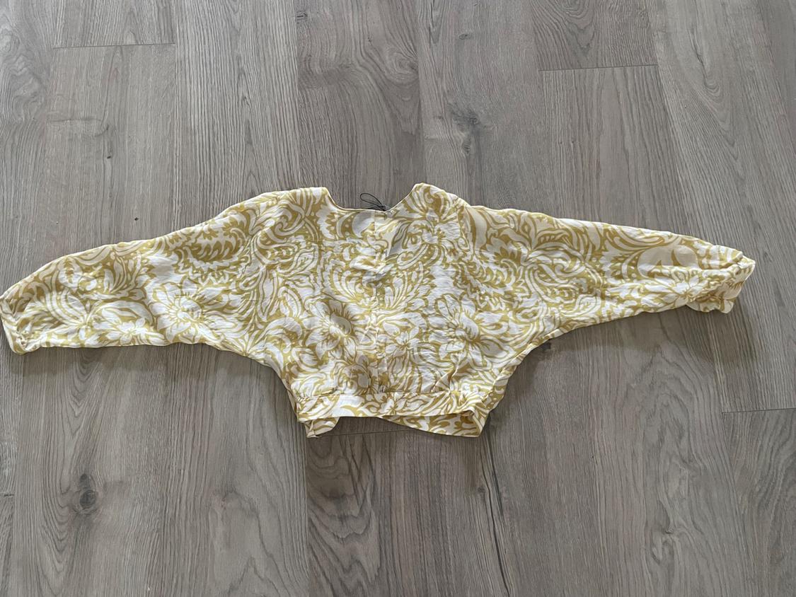 Long-sleeved blouse with yellow floral print on a white background, spread out on a gray wooden floor.