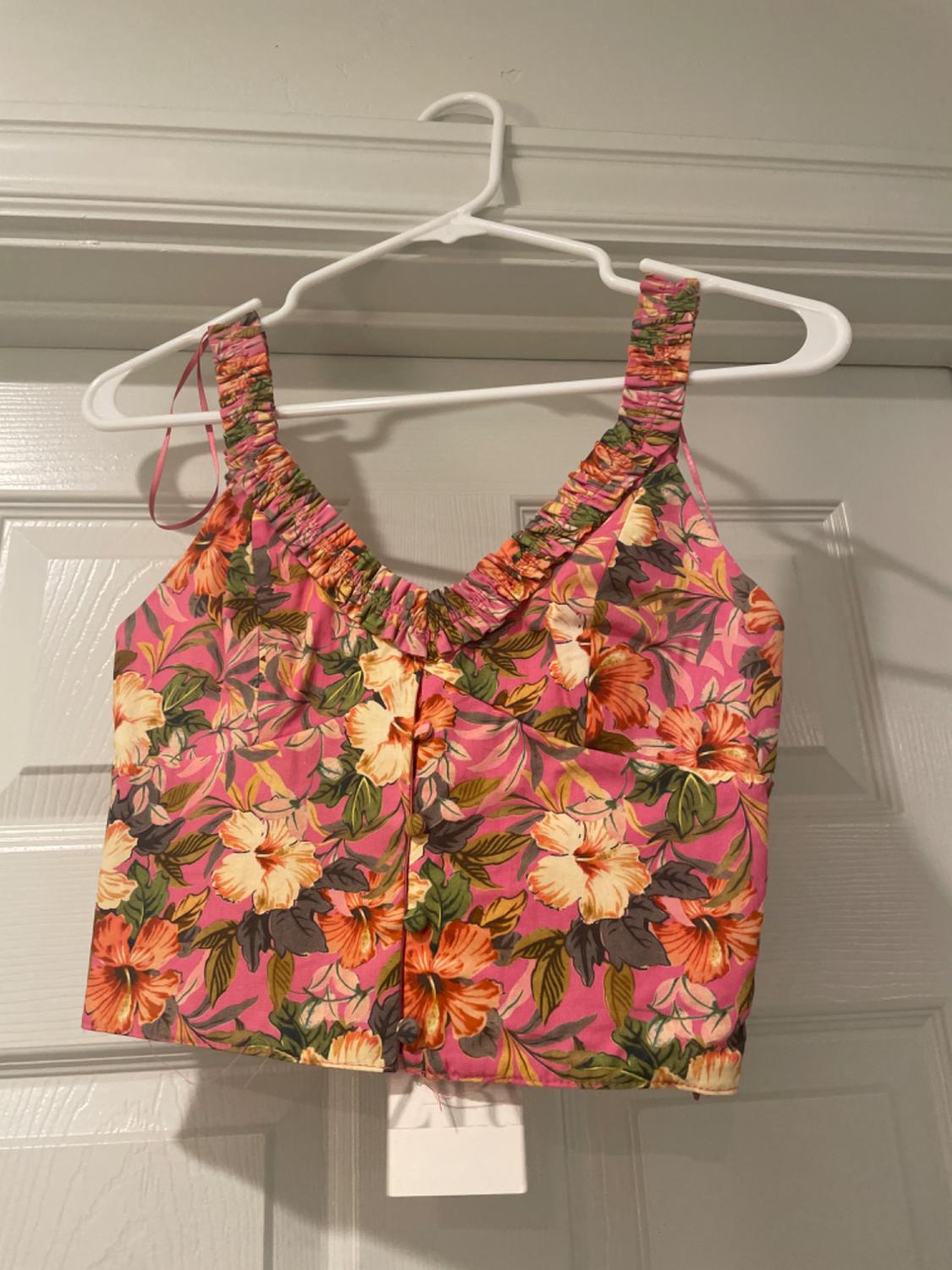 Strappy top with tropical floral print in shades of pink, orange and green, hanging on a white hanger on a white door.