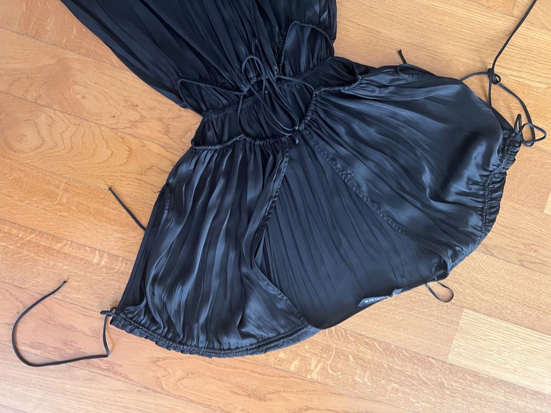 Black pleated garment with a satin finish featuring cut-outs and laces, laid out on a light wood floor.