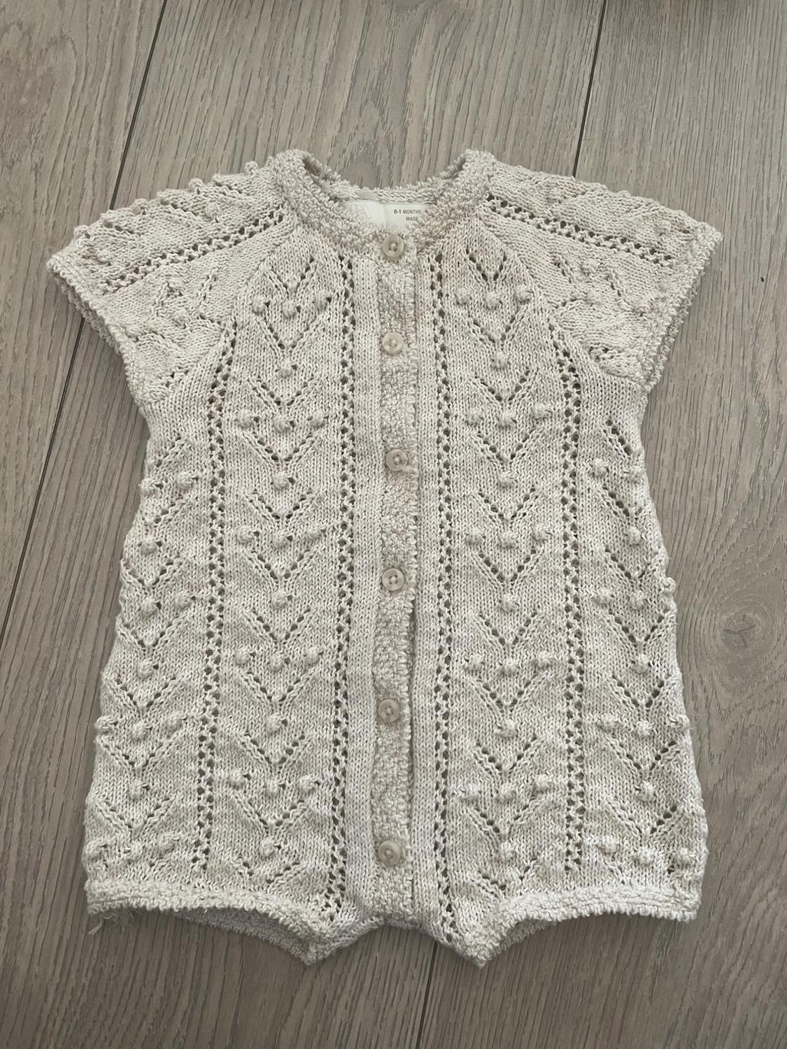 Knitted baby romper in beige with front buttons, on a light wooden floor. Knitted design with openwork details.
