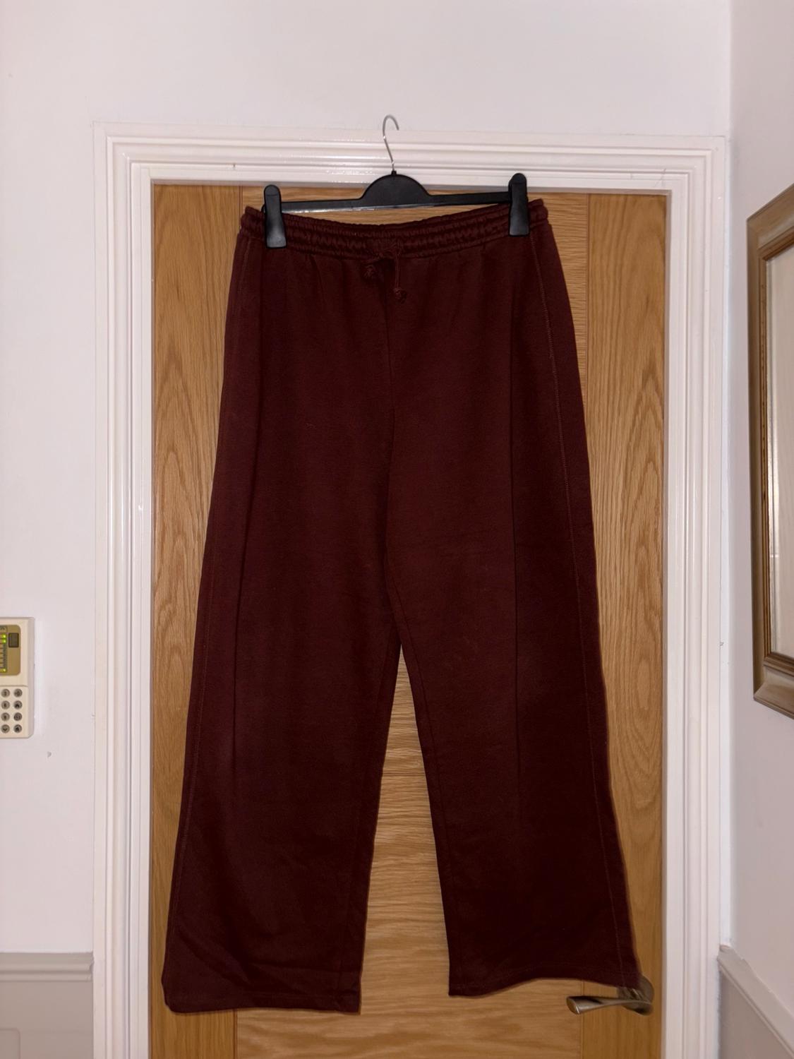 Burgundy tracksuit bottoms with wide legs hanging on a light wooden door, showing a casual, relaxed style.