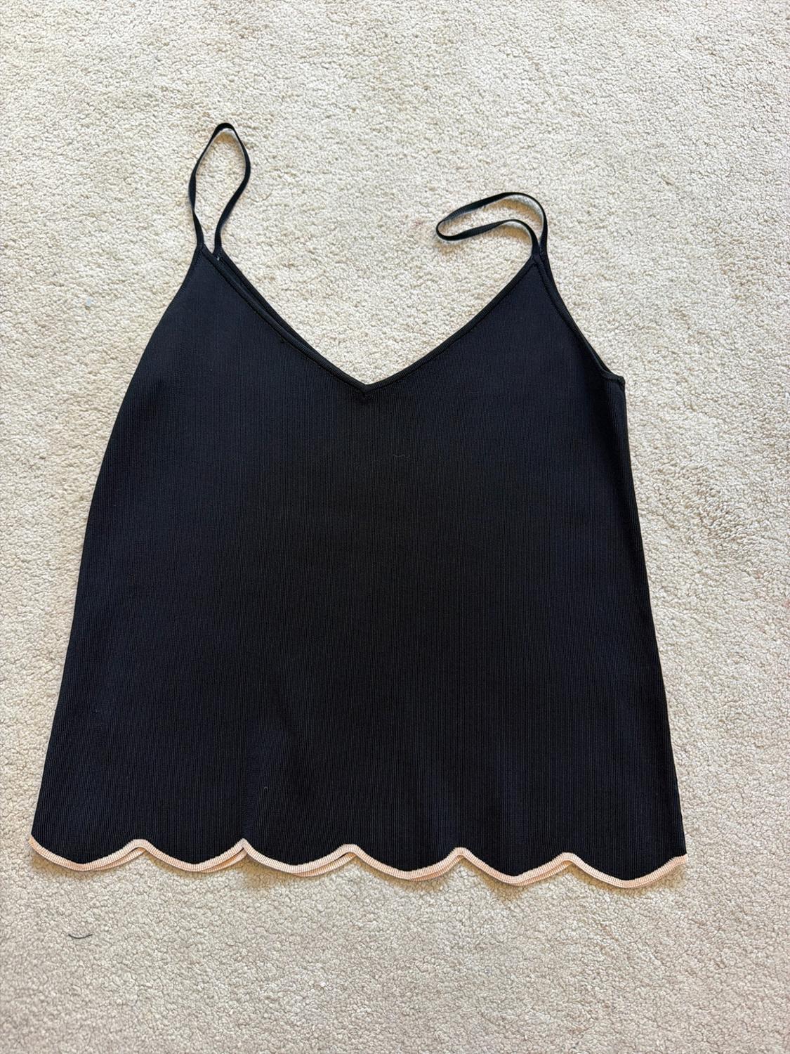 Black knitted camisole top with V-neck, spaghetti straps and a scalloped hem with light-coloured trim, on a textured rug.