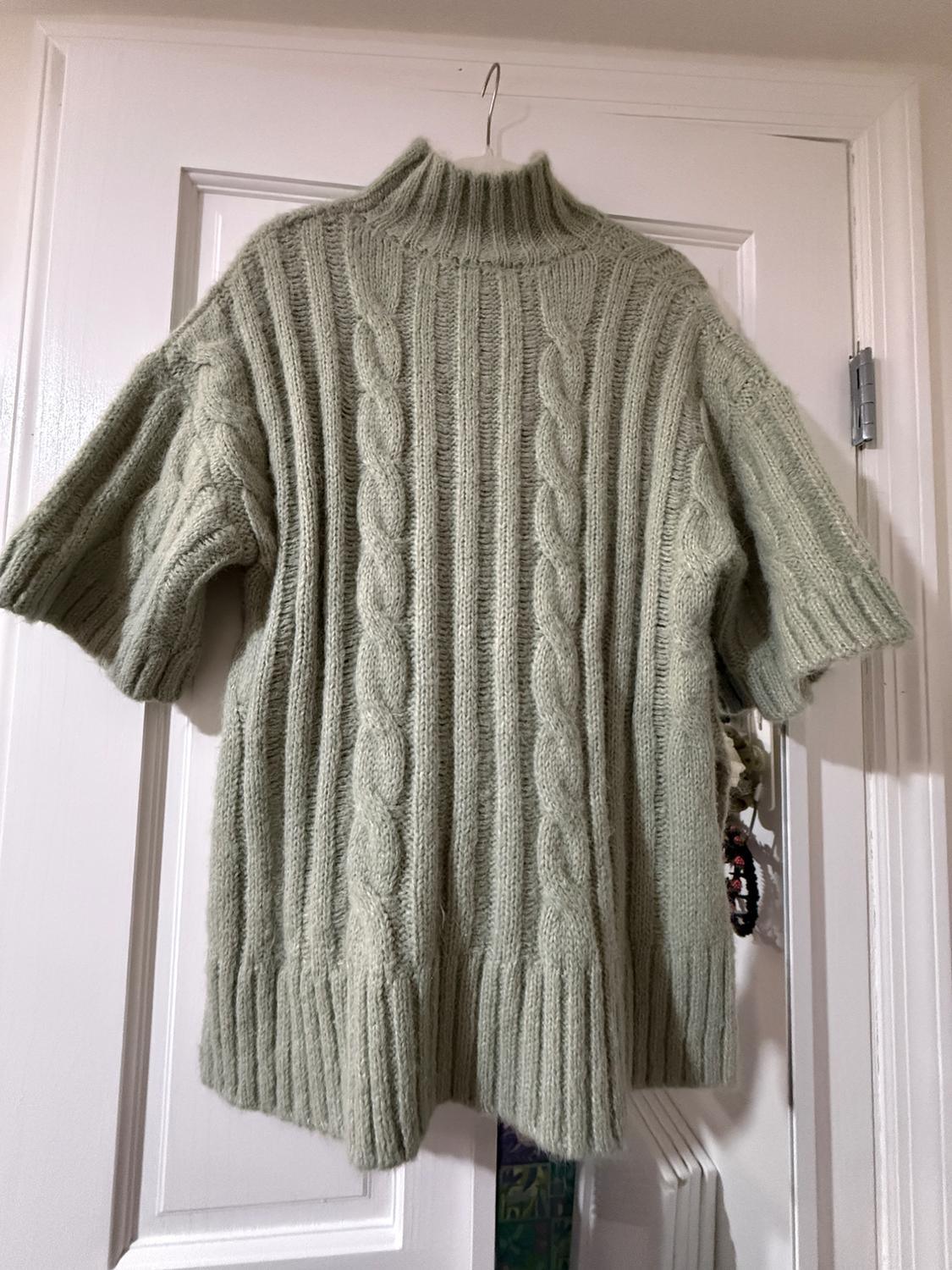 Light green chunky knit jumper with a high neck and short sleeves, hanging on a white door.