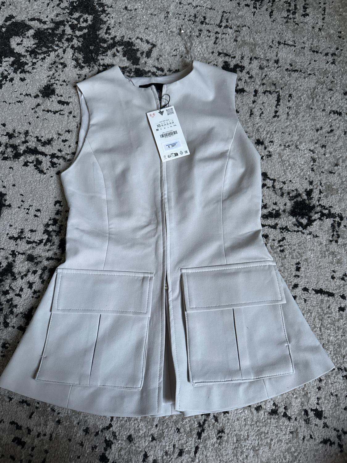 Light grey sleeveless gilet with large pockets and a swing tag on a black and white abstract design rug.