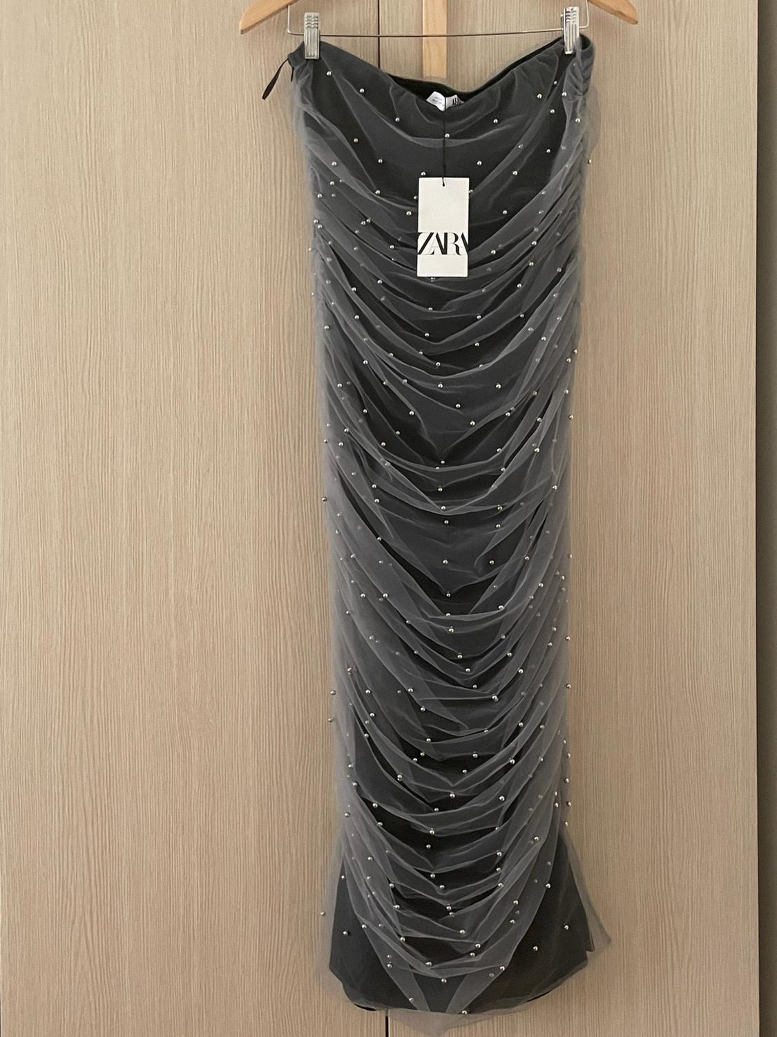 Black ruched tulle dress with silver pearls hanging on a hanger against a wooden background. White tag with the text ZARA.