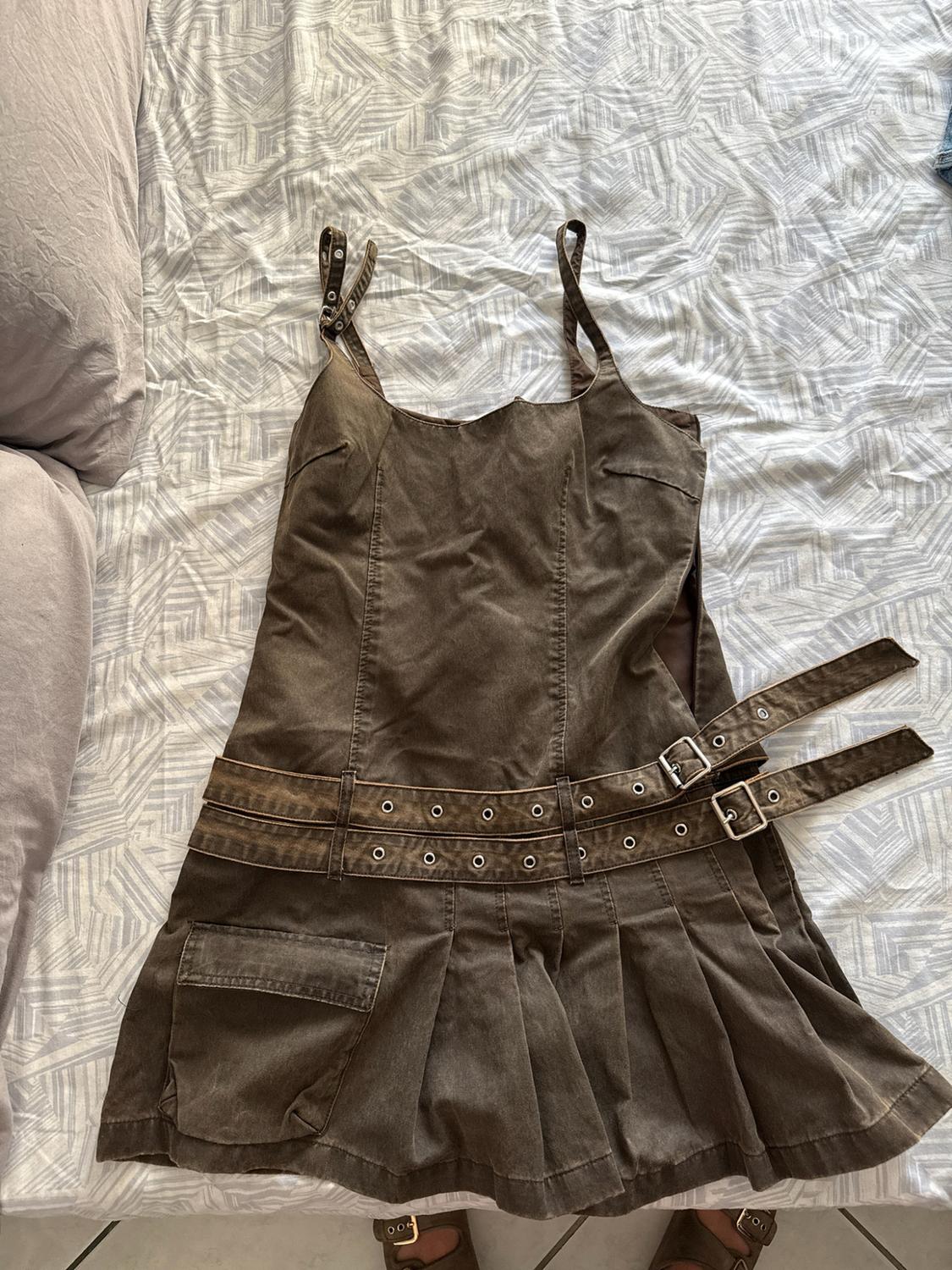 Short brown dress with a pleated skirt, shoulder straps and a double belt with buckles, photographed on a bed with patterned sheets.